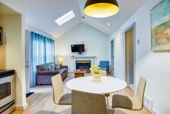 A modern living room with a fireplace, a television mounted above it, and a dining area with a round table and chairs.