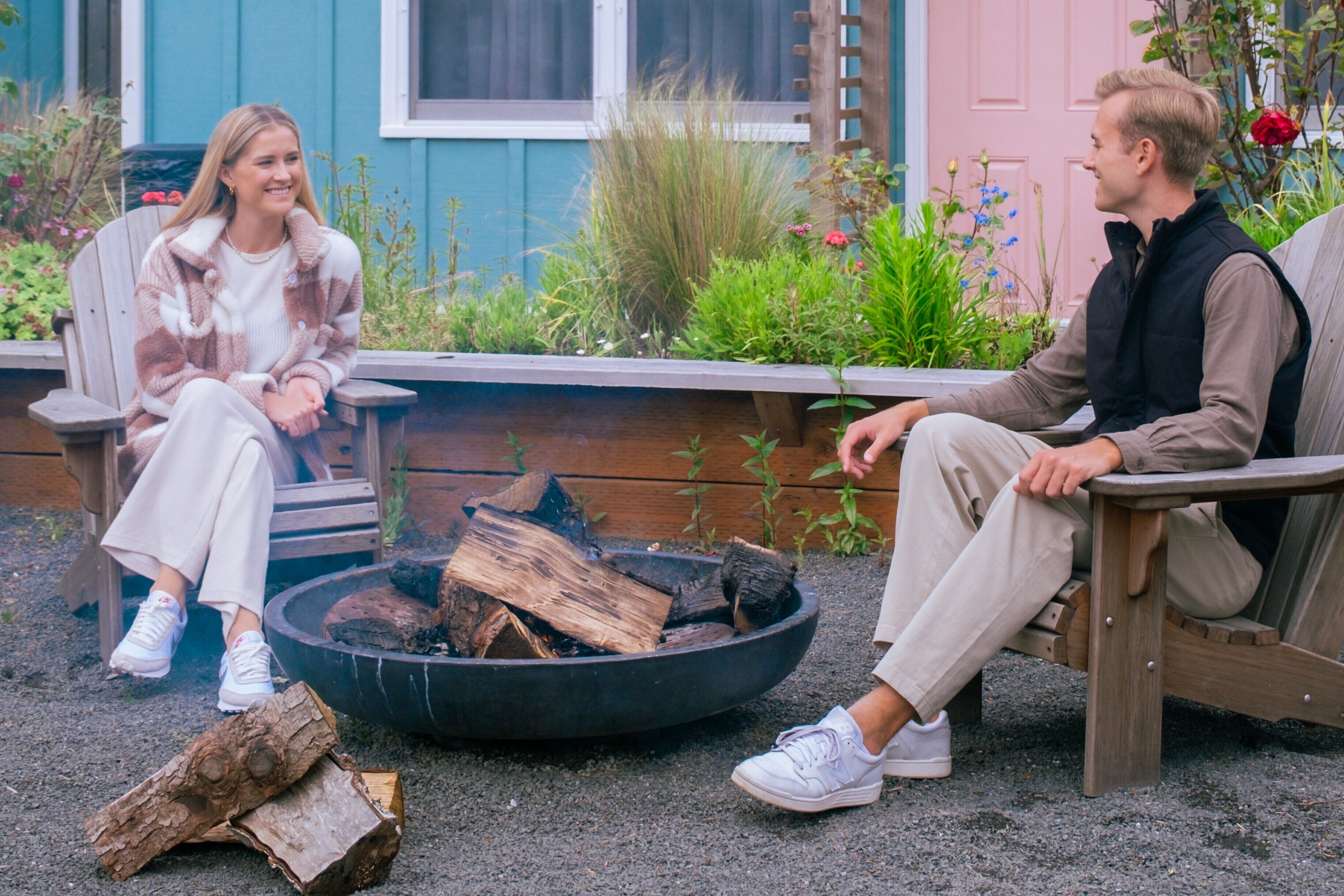 Two people sitting on wooden benches near a fire pit with logs.