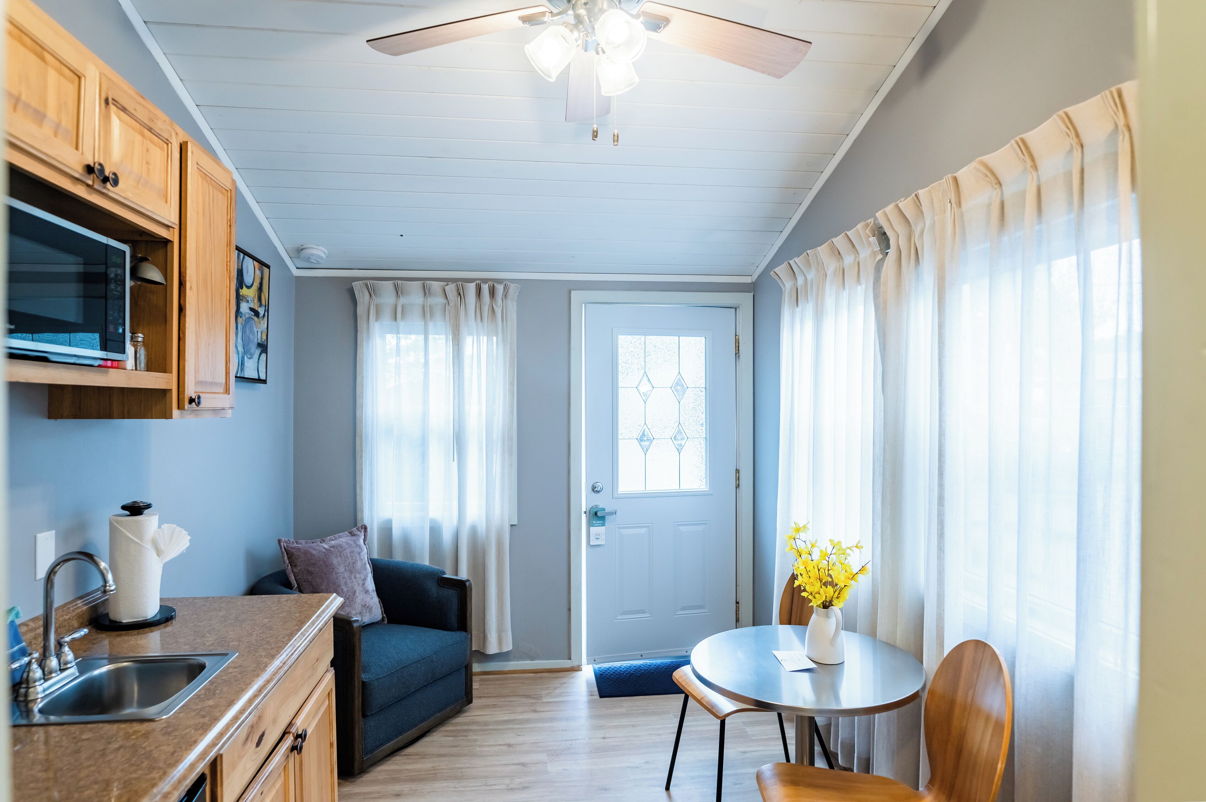 A compact kitchen with a microwave, sink, and wooden cabinets. There is a dining area with a table, chairs, and a vase of flowers.