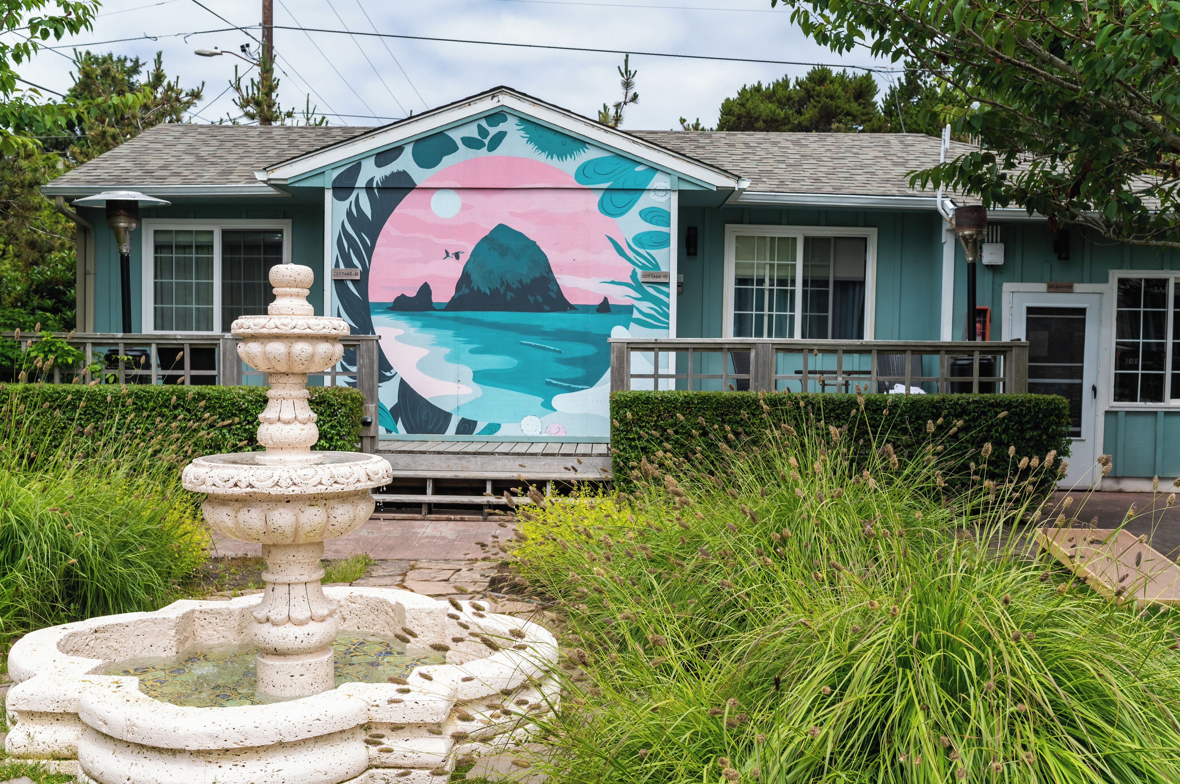Un edificio con un mural de un atardecer sobre el agua, una fuente de piedra y abundante vegetación.
