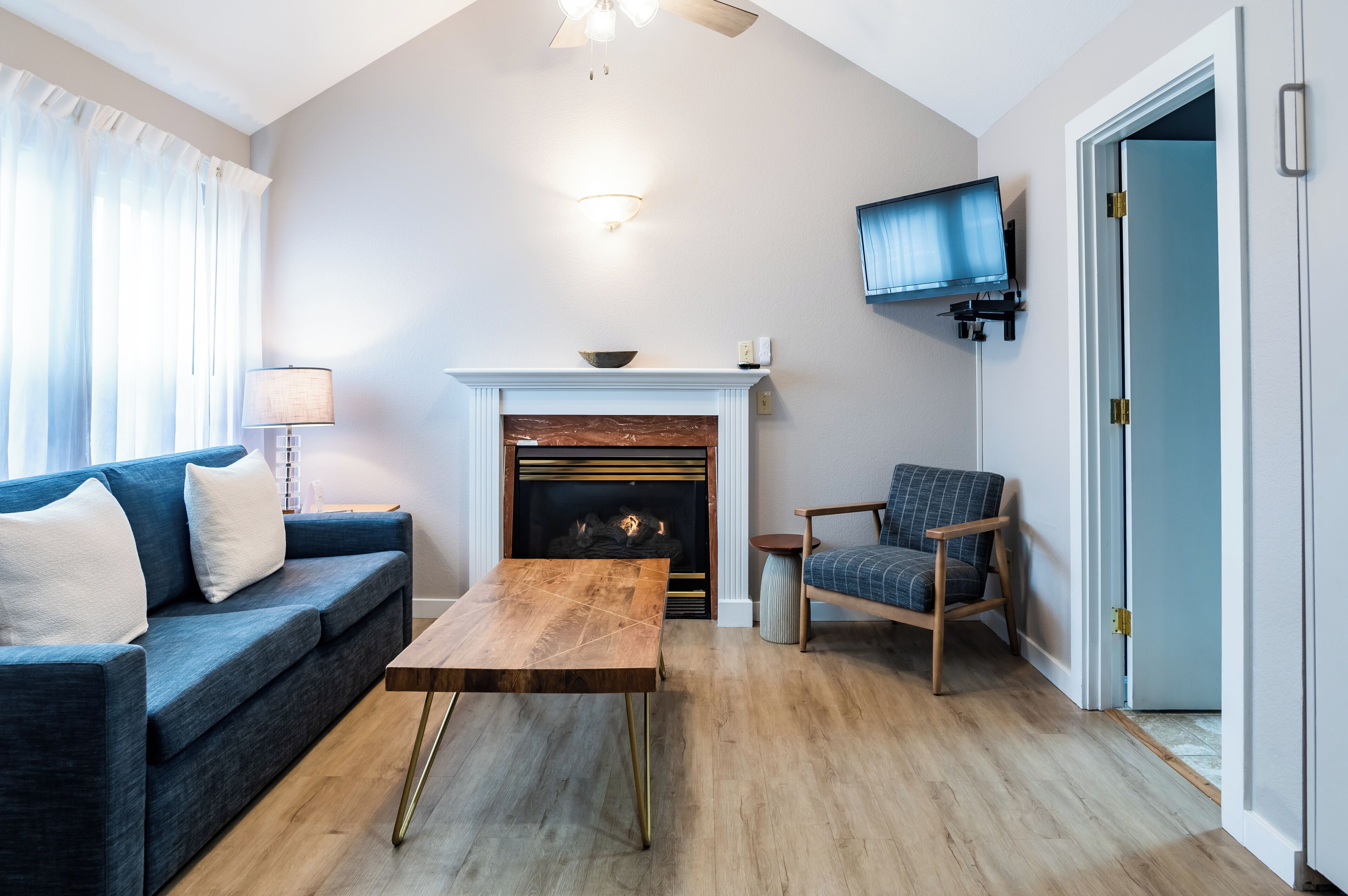 Una sala de estar con un sofá azul, una mesa de centro de madera, una chimenea, un televisor montado en la pared y una puerta que da a otra habitación.
