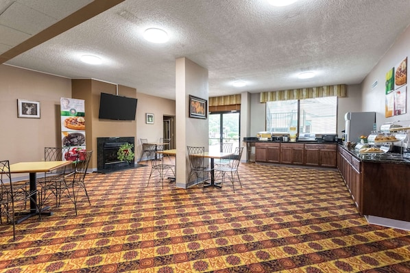 Um saguão de hotel com carpete estampado, lareira, televisão fixada na parede e uma área de cozinha com buffet.