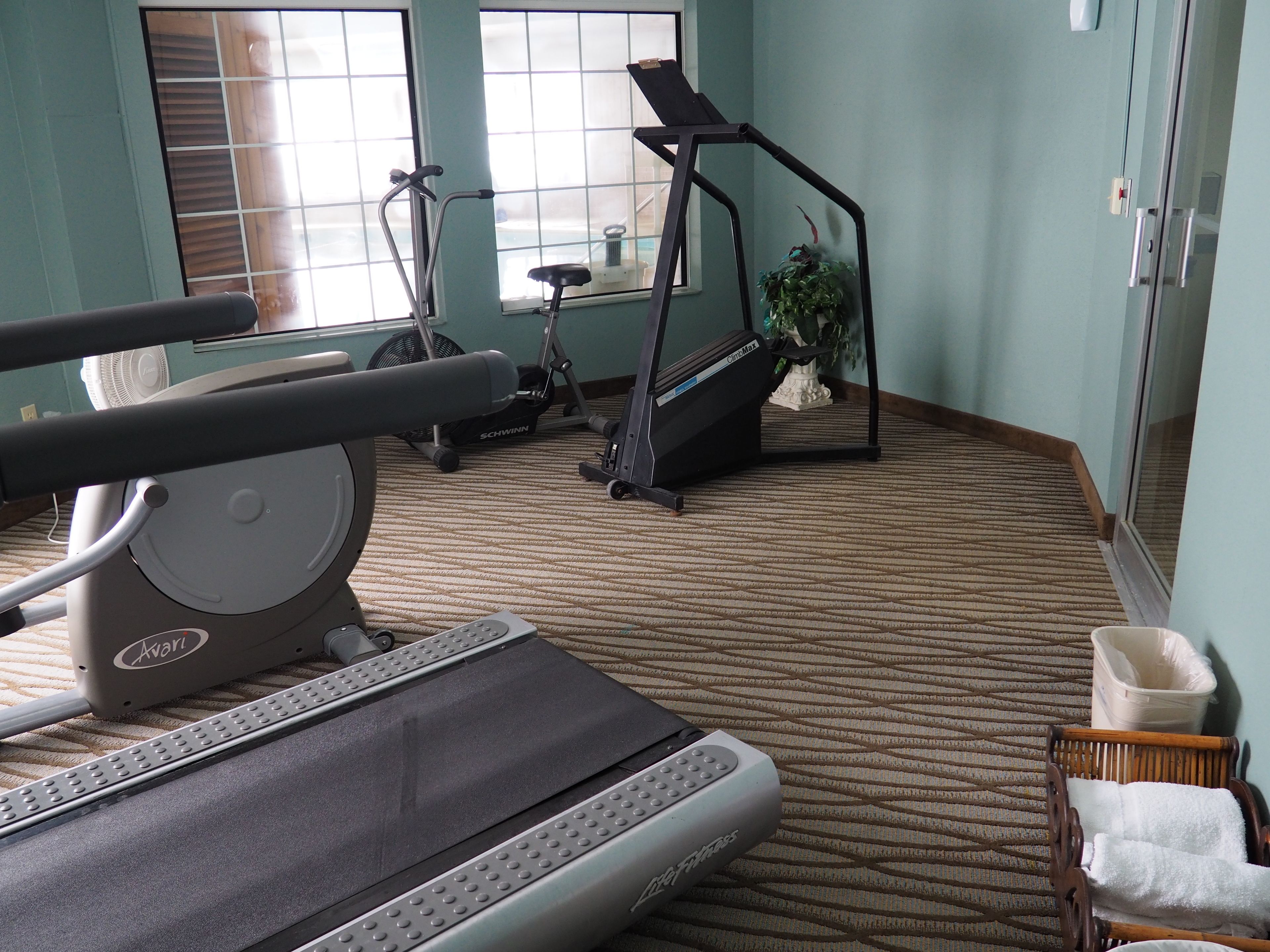 A hotel gym with exercise equipment, a carpeted floor, and a window with blinds.