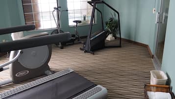 Gimnasio de hotel con equipamiento de ejercicio, piso alfombrado y una ventana con persianas.