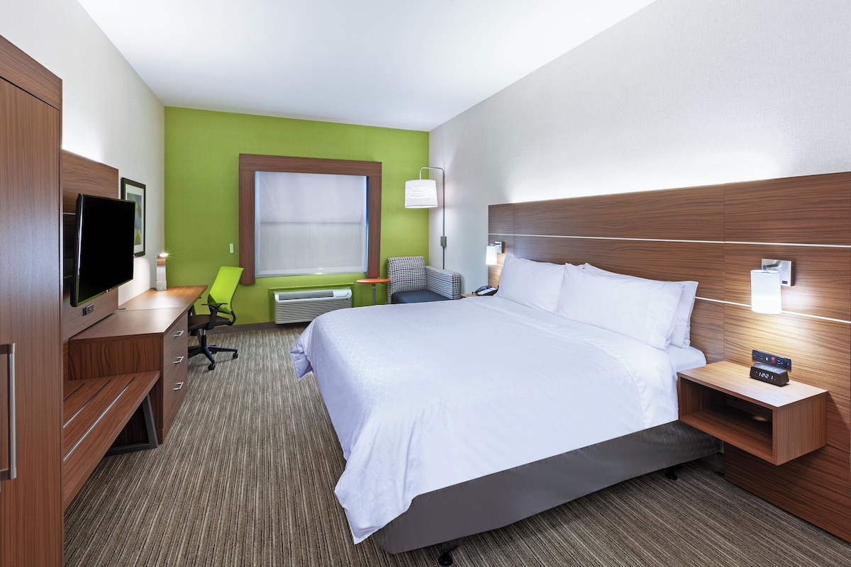 Une chambre d’hôtel avec un grand lit, un bureau avec une chaise, une télévision et une fenêtre avec des stores.