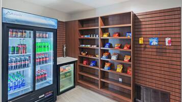 Una máquina expendedora con diversas bebidas y aperitivos, un refrigerador pequeño y estantes de madera llenos de bocadillos.