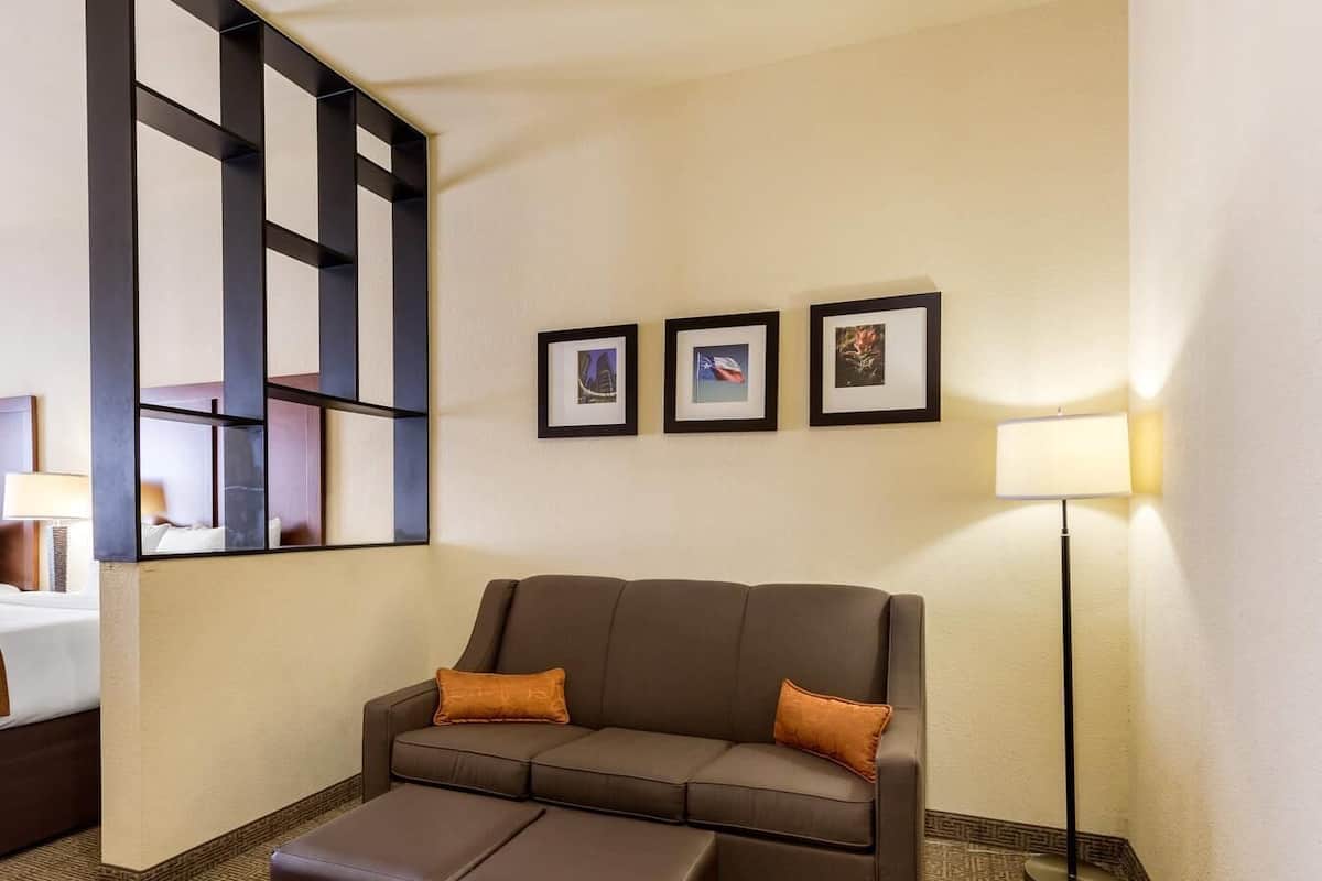 A hotel room with a brown sofa, a matching ottoman, a floor lamp, and three framed pictures on the wall.