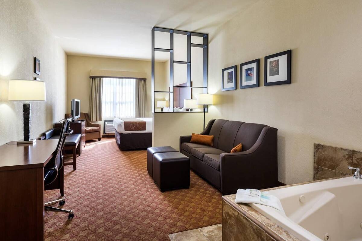 A hotel room with a bed, a sofa, a desk with a computer, a TV, a bathroom with a bathtub, and framed pictures on the wall.