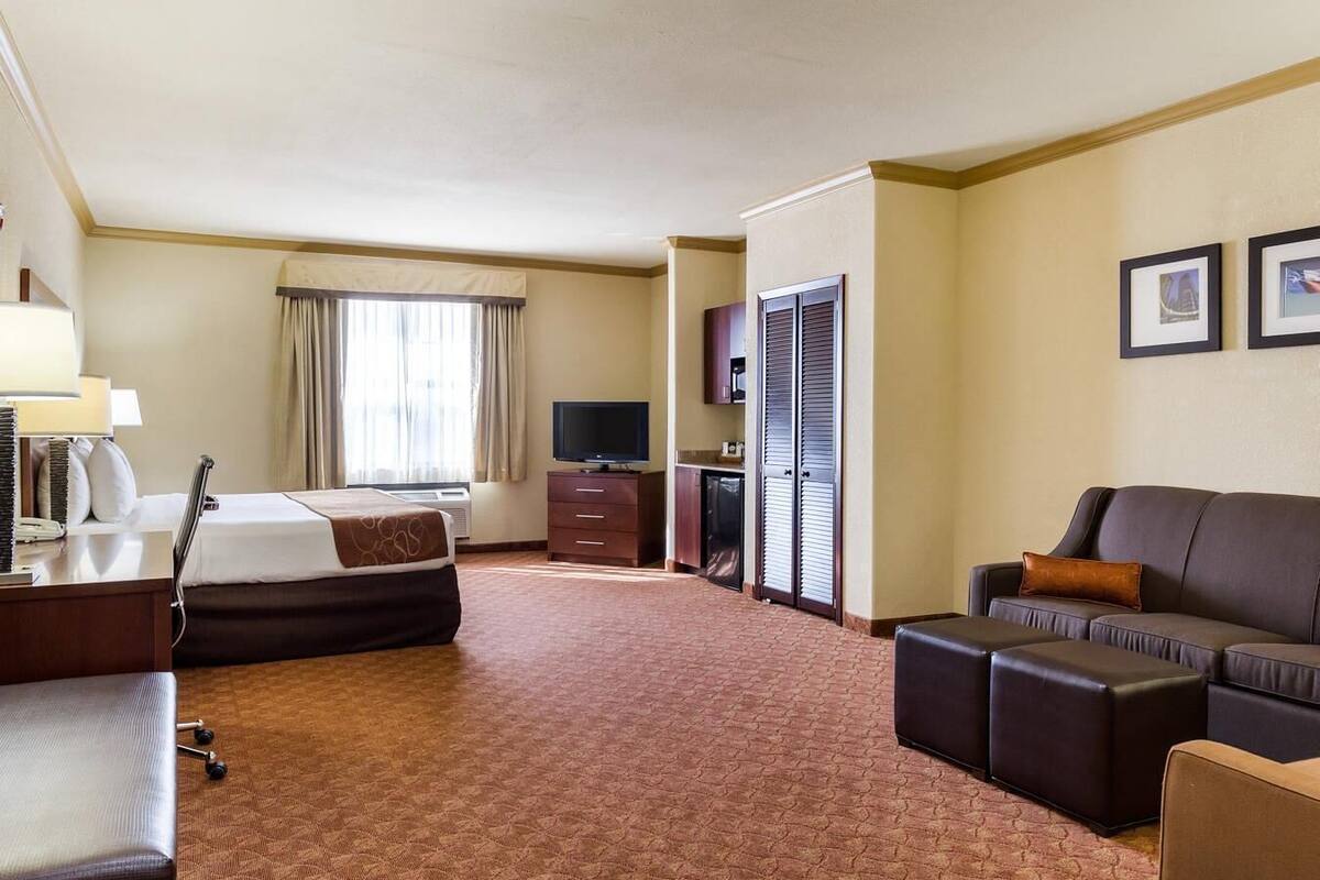 A hotel room with a bed, a desk with a lamp, a television, a sofa, and a large window with curtains.