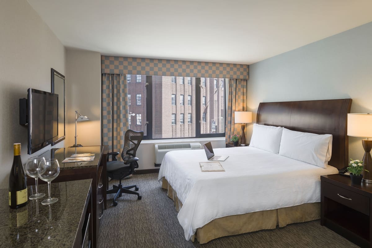 Habitación de hotel con cama, escritorio con laptop, televisión, silla, ventana con vista a edificios y lámpara.