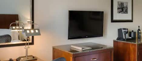 A hotel room with a bed, a desk with a lamp, a chair, a TV, a dresser, and a framed picture on the wall.