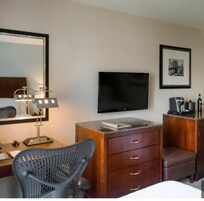 A hotel room with a bed, a desk with a lamp, a chair, a TV, a dresser, and a framed picture on the wall.