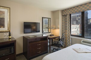 A hotel room with a bed, a desk, a chair, a television, a minibar, and a window with a city view.