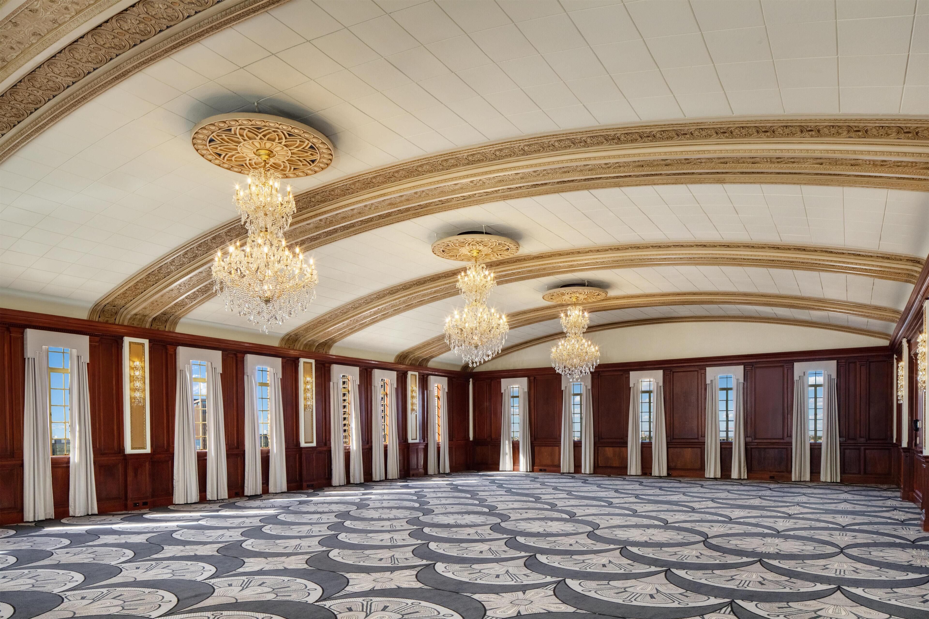 Um salão espaçoso e ricamente decorado, com piso estampado, janelas altas com cortinas e várias luminárias de cristal.