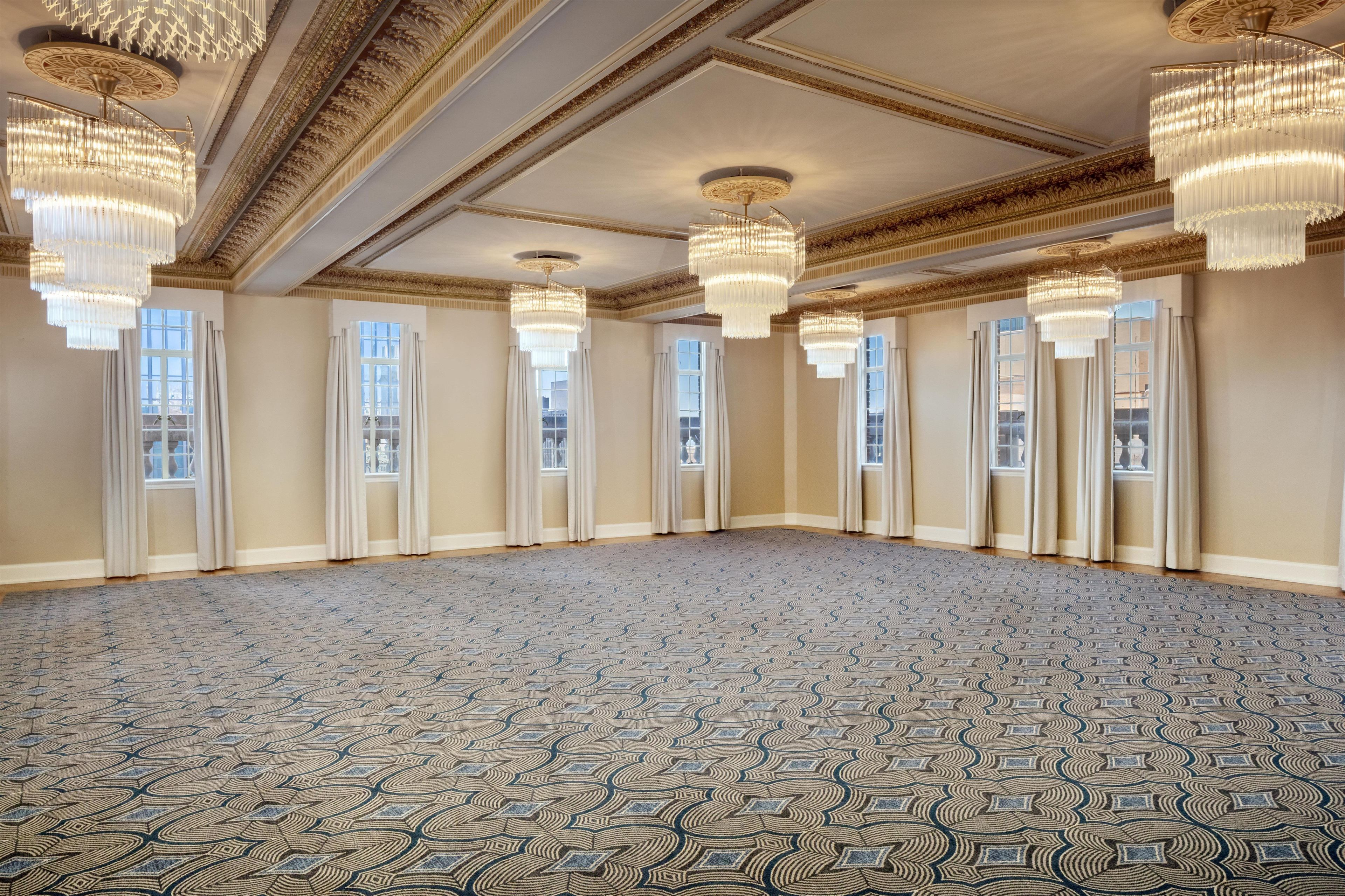A spacious room with a patterned carpet, large windows with curtains, and multiple chandeliers.