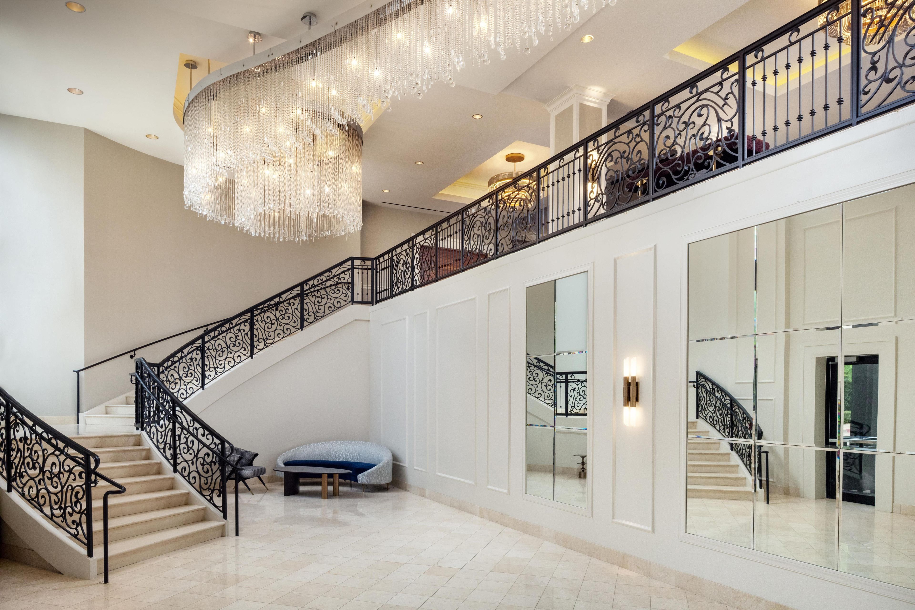 A grand staircase with ornate black railings, a chandelier, and a seating area with a blue bench.