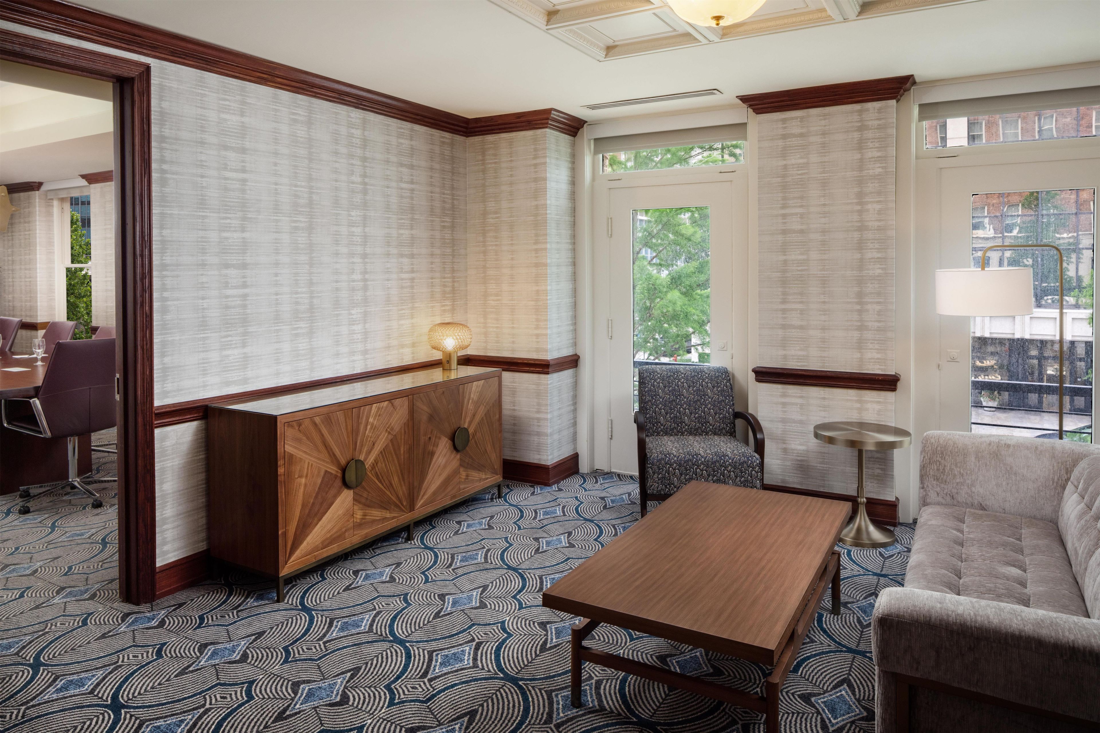 A hotel room with a wooden console table, patterned carpet, a sofa, and a chair.