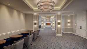 Ein Hotelkorridor mit einem gemusterten Teppich, einer Sitzecke mit runden Tischen und Stühlen sowie einem großen Kronleuchter.