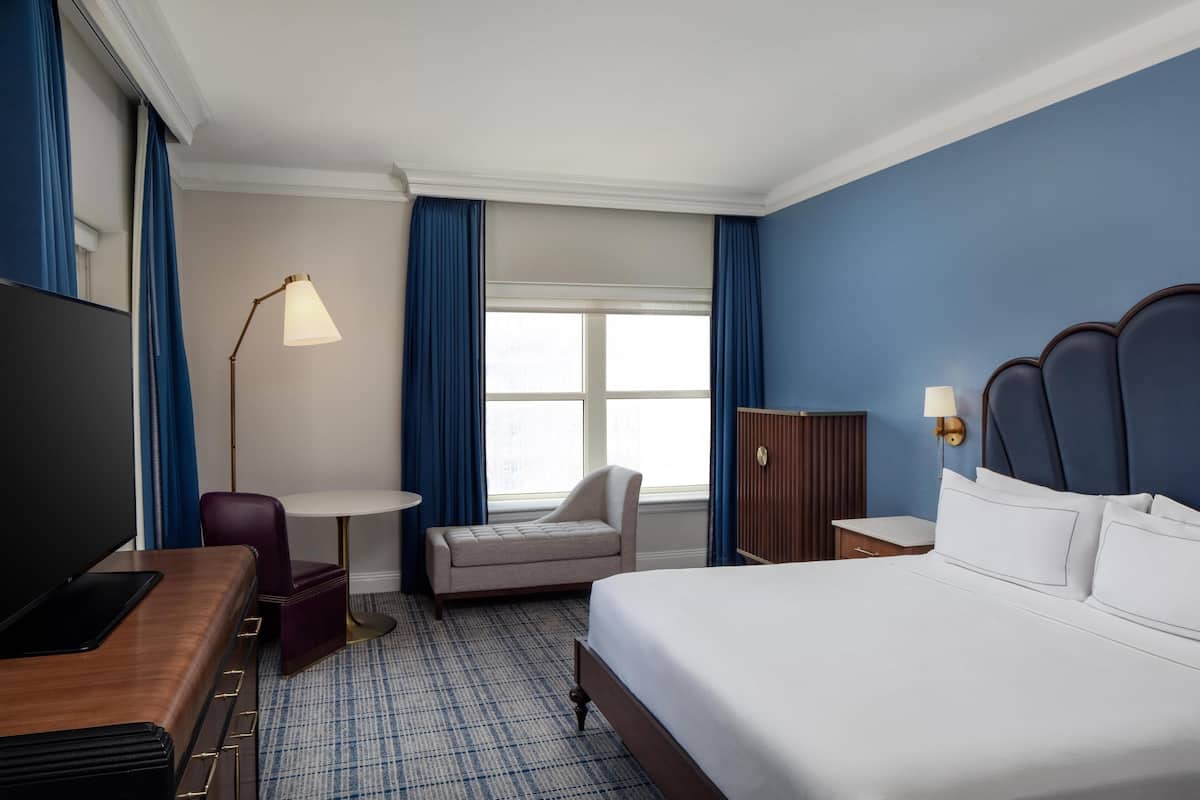 Ein Hotelzimmer mit einem großen Bett, einem Schreibtisch, einem Flachbildfernseher, einem Sessel, einem kleinen Tisch und einer Lampe.