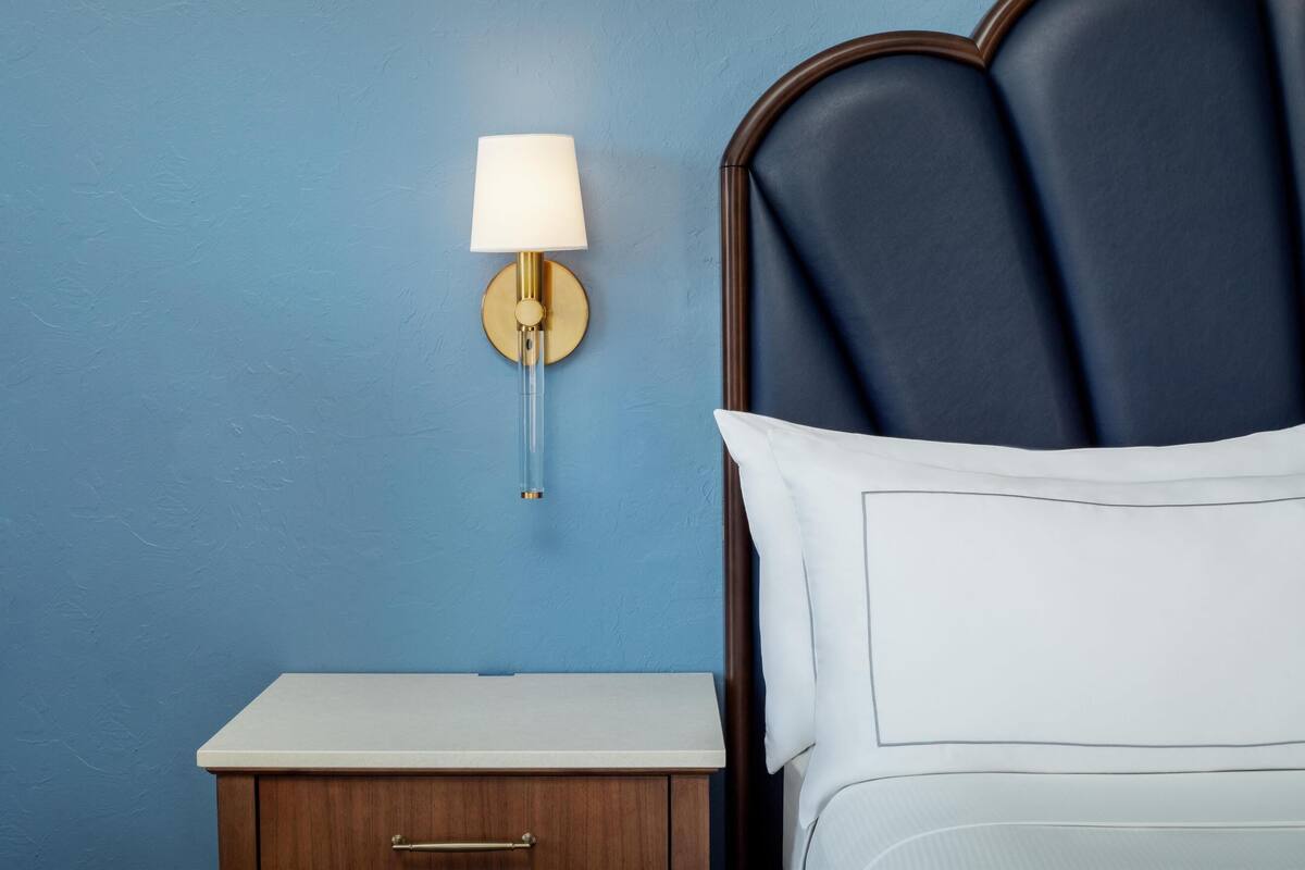Ein ordentlich bezogenes Bett mit weißem Kopfteil und einem Nachttisch mit Lampe daneben.
