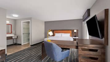 Habitación de hotel con cama, escritorio, silla, televisor y baño con ducha.