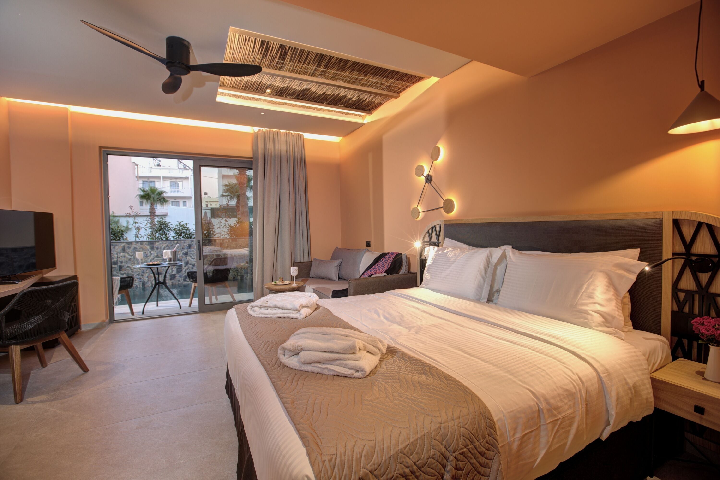 Ein modernes Hotelzimmer mit einem großen Bett, einem Balkon mit Tisch und Stühlen, einem Deckenventilator und einem Fernseher.