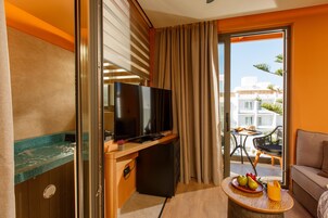 A hotel room with a TV, a small fridge, a balcony with a table and chairs, and a sofa.