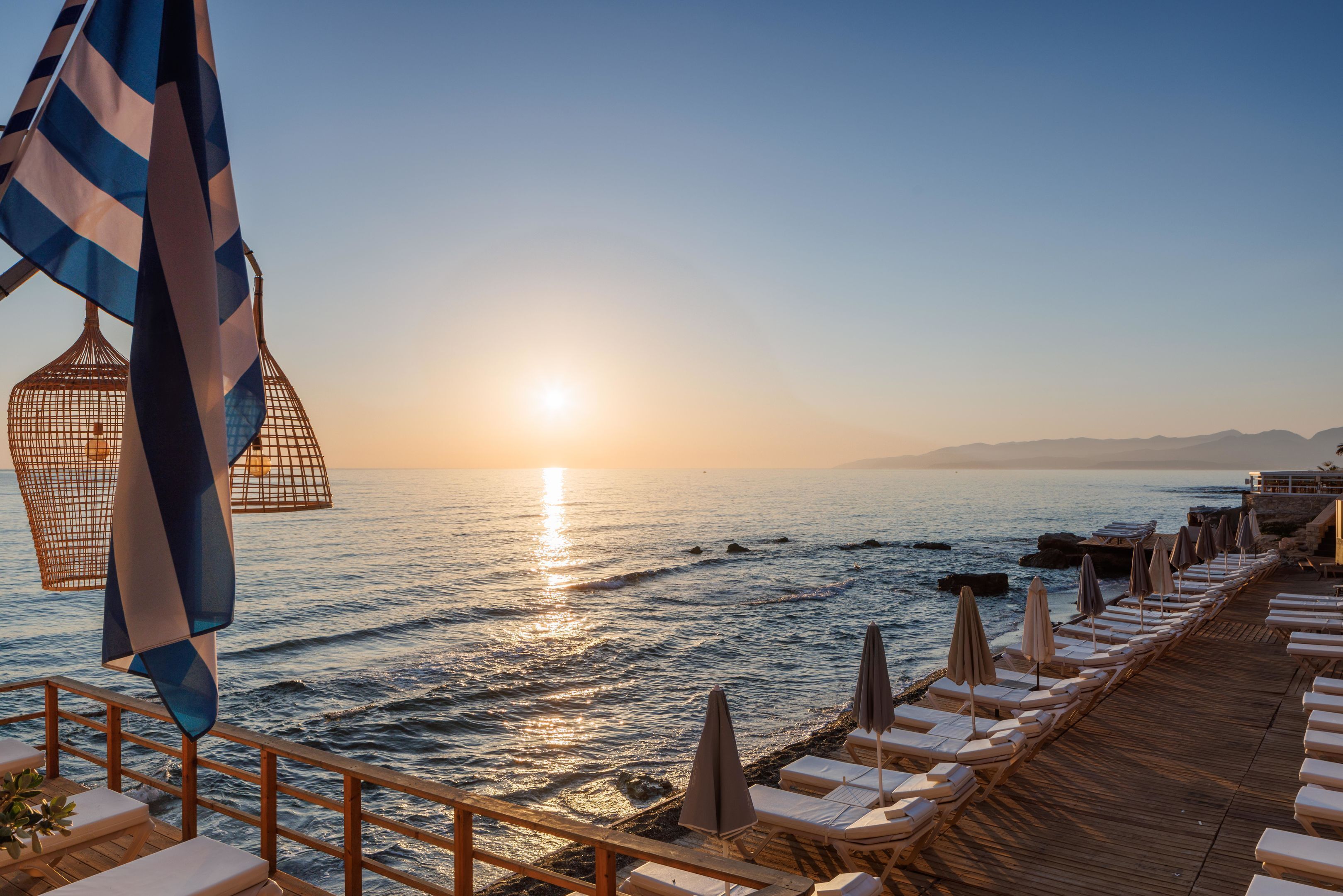 A wooden deck with lounge chairs and umbrellas overlooking a beach at sunset.