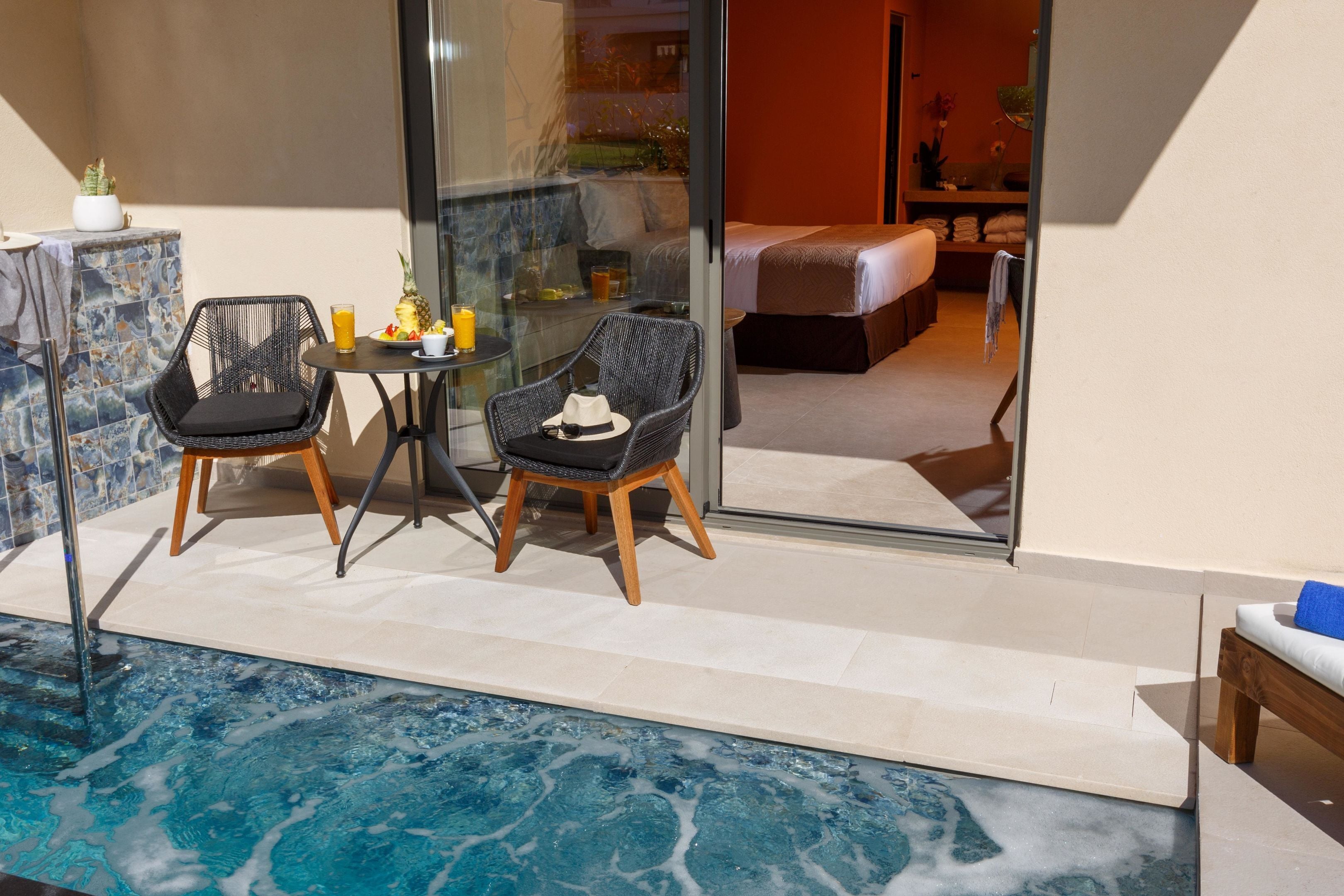 A poolside area with two wicker chairs, a small table with a drink and fruit, and a glass of orange juice.