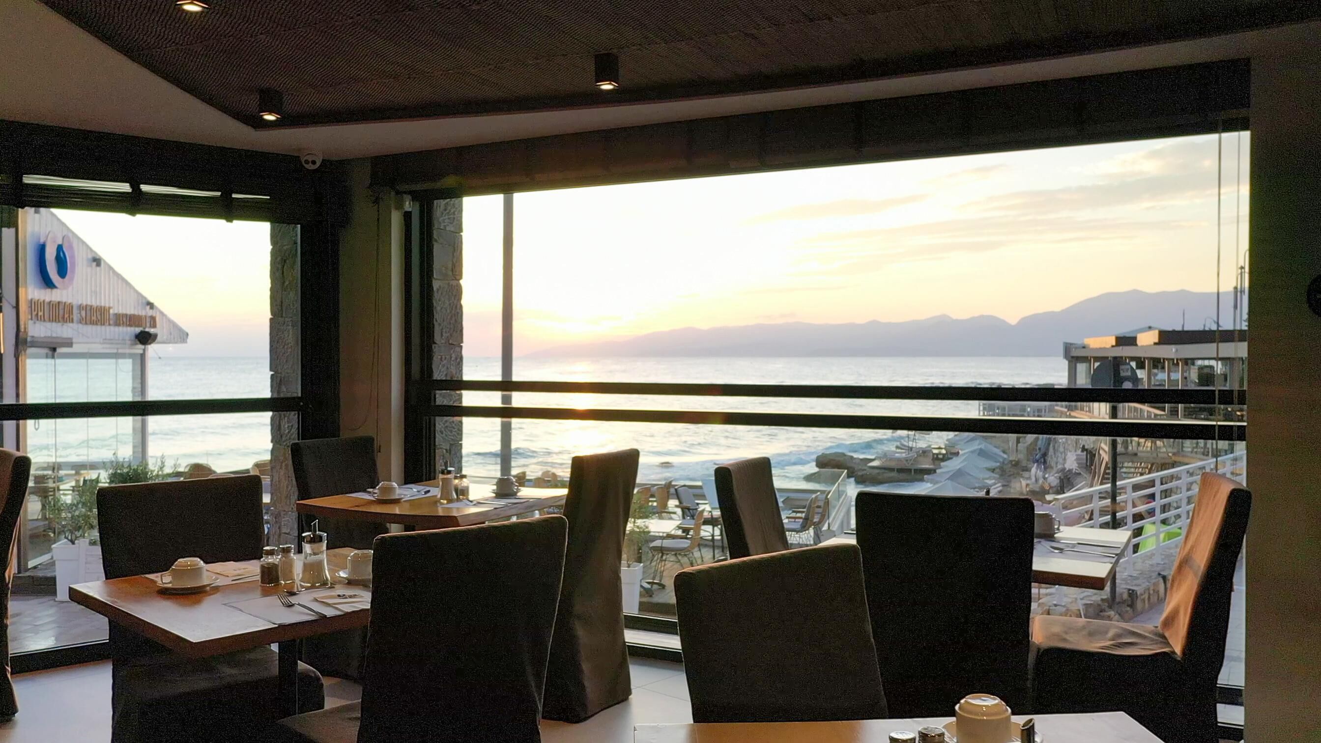 A restaurant with a view of the sea and mountains. Tables are set with plates, cups, and cutlery.