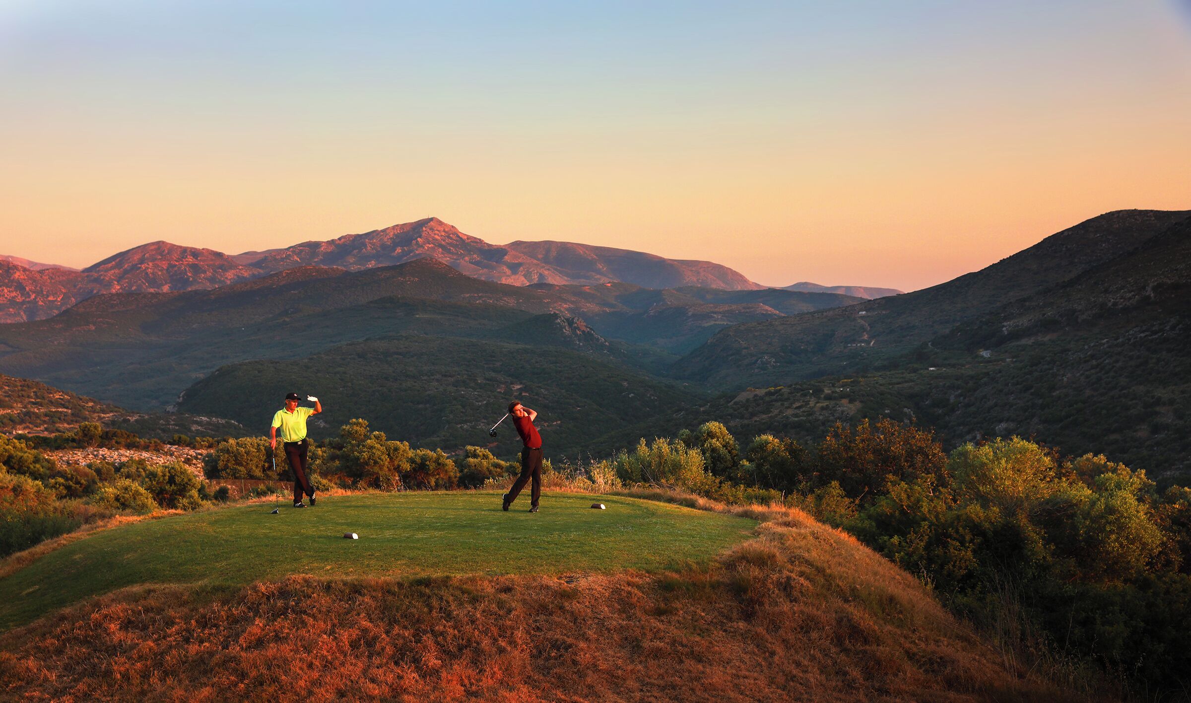 A golf course with two players in a valley surrounded by mountains.