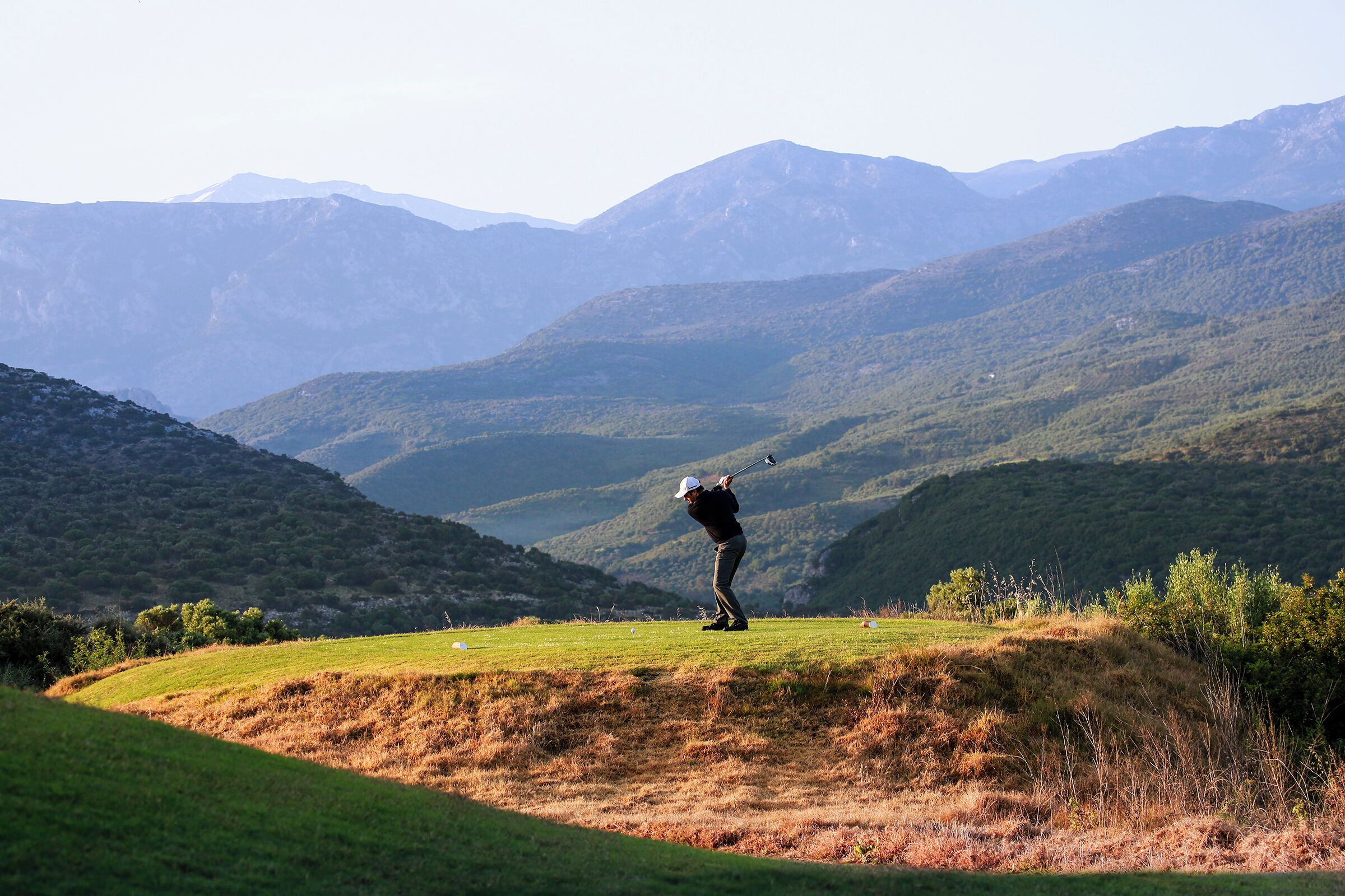 A golfer on a grassy hill with a mountainous backdrop.