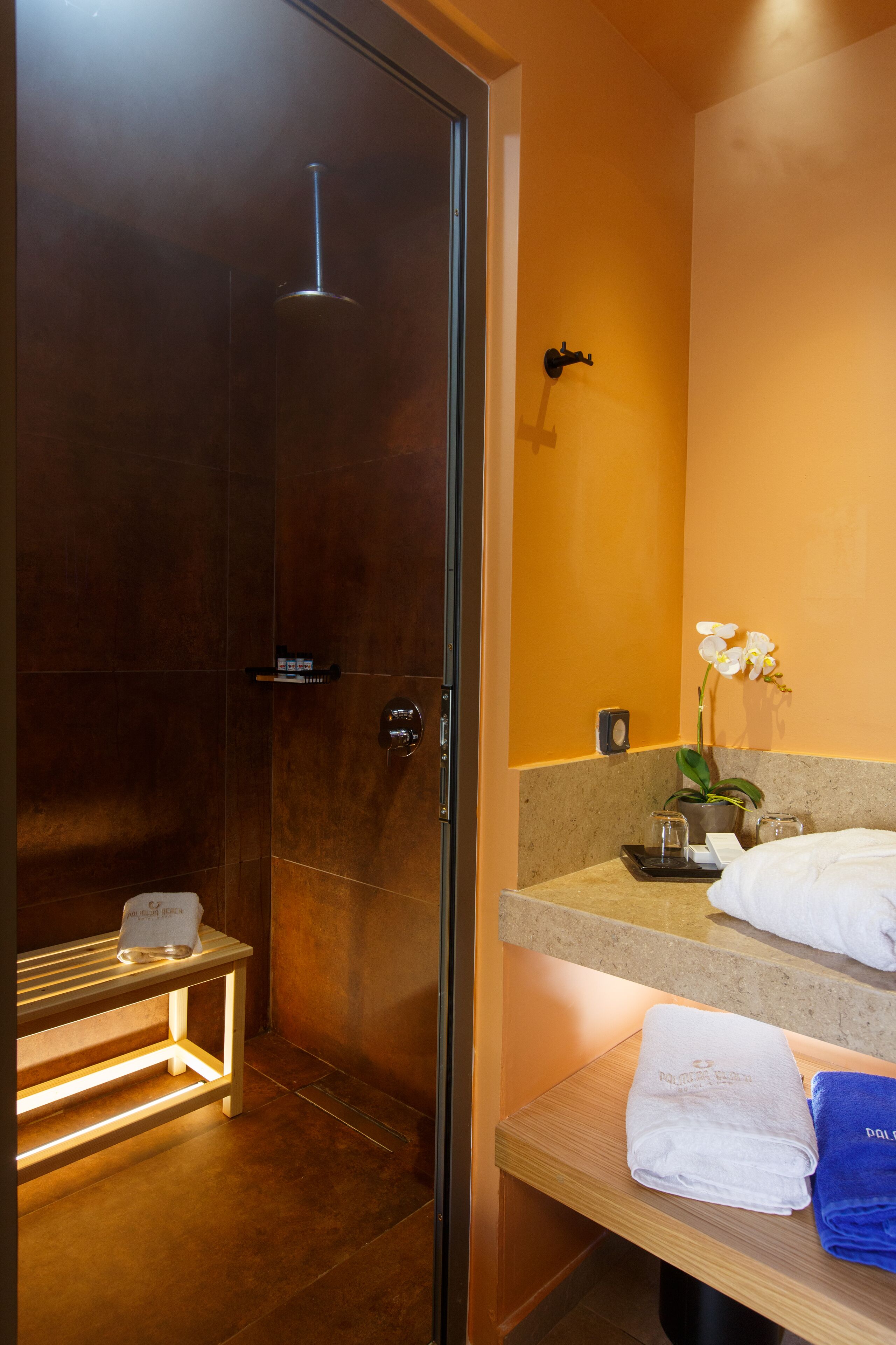 Une salle de bain moderne avec une douche à parois de verre, un banc en bois, un lavabo avec un miroir et une orchidée en pot.