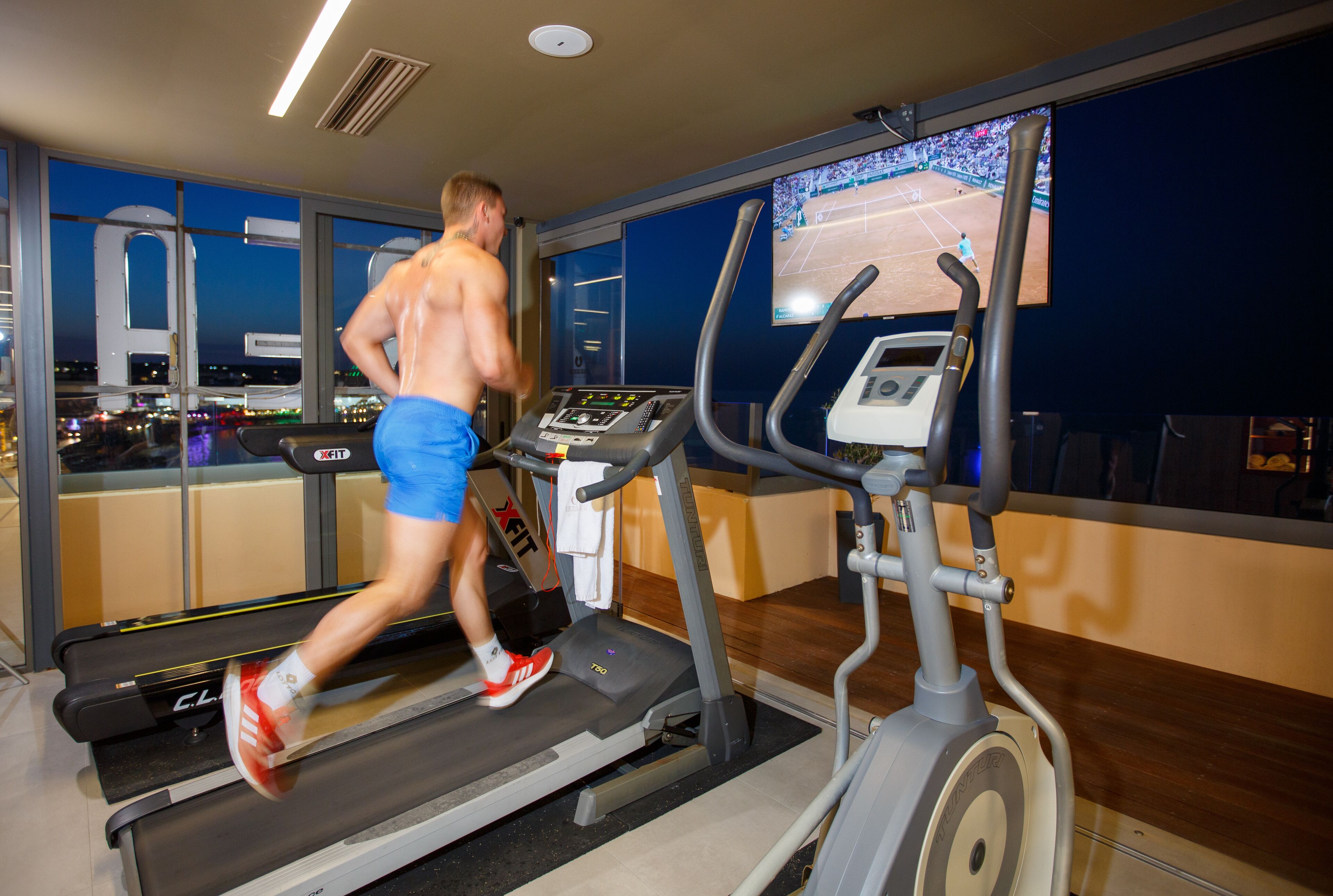 A man running on a treadmill in a gym with a large window and a television displaying a tennis match.