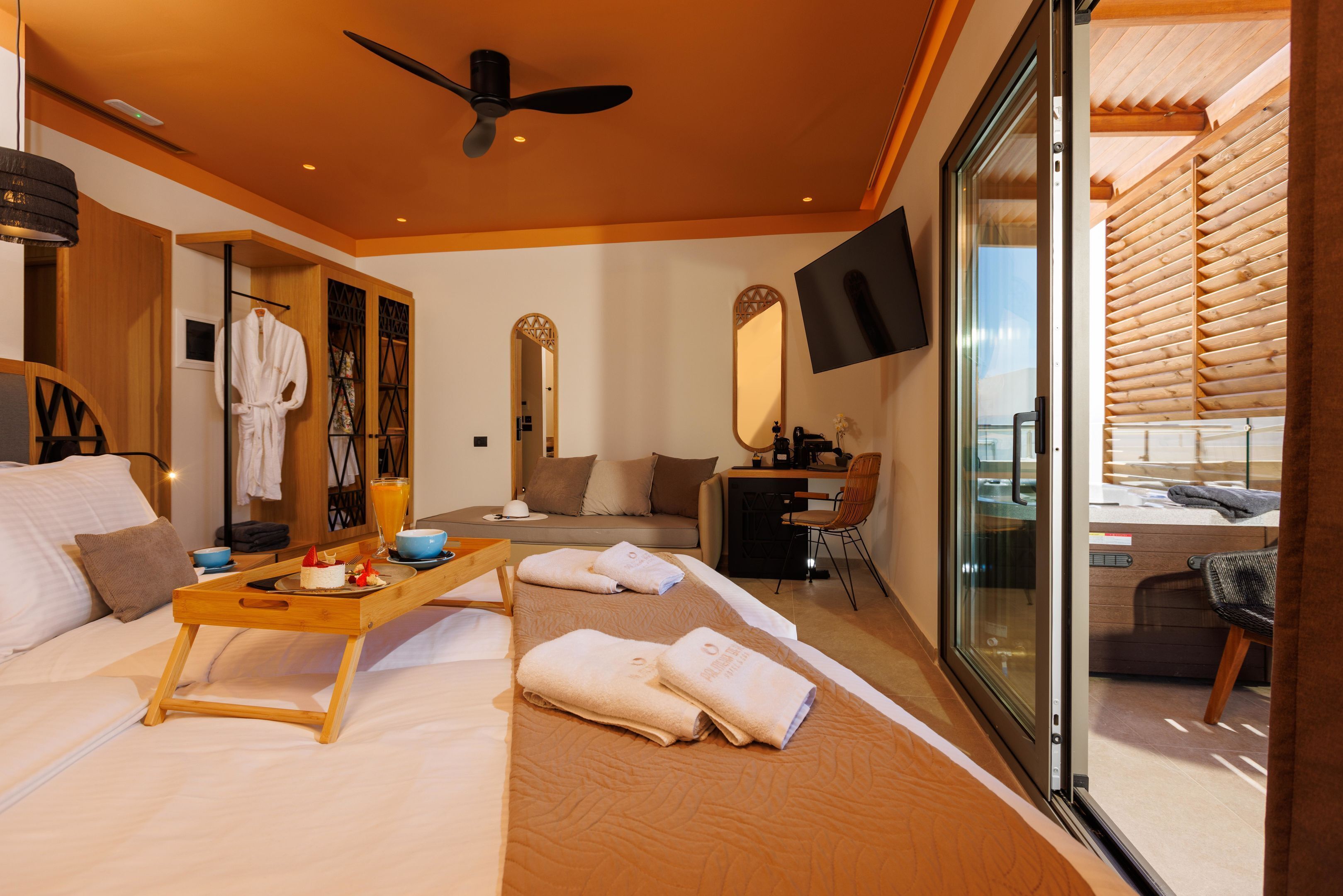 A modern hotel room with a bed, a wooden side table with a tray, a sofa, a desk, and a large mirror.