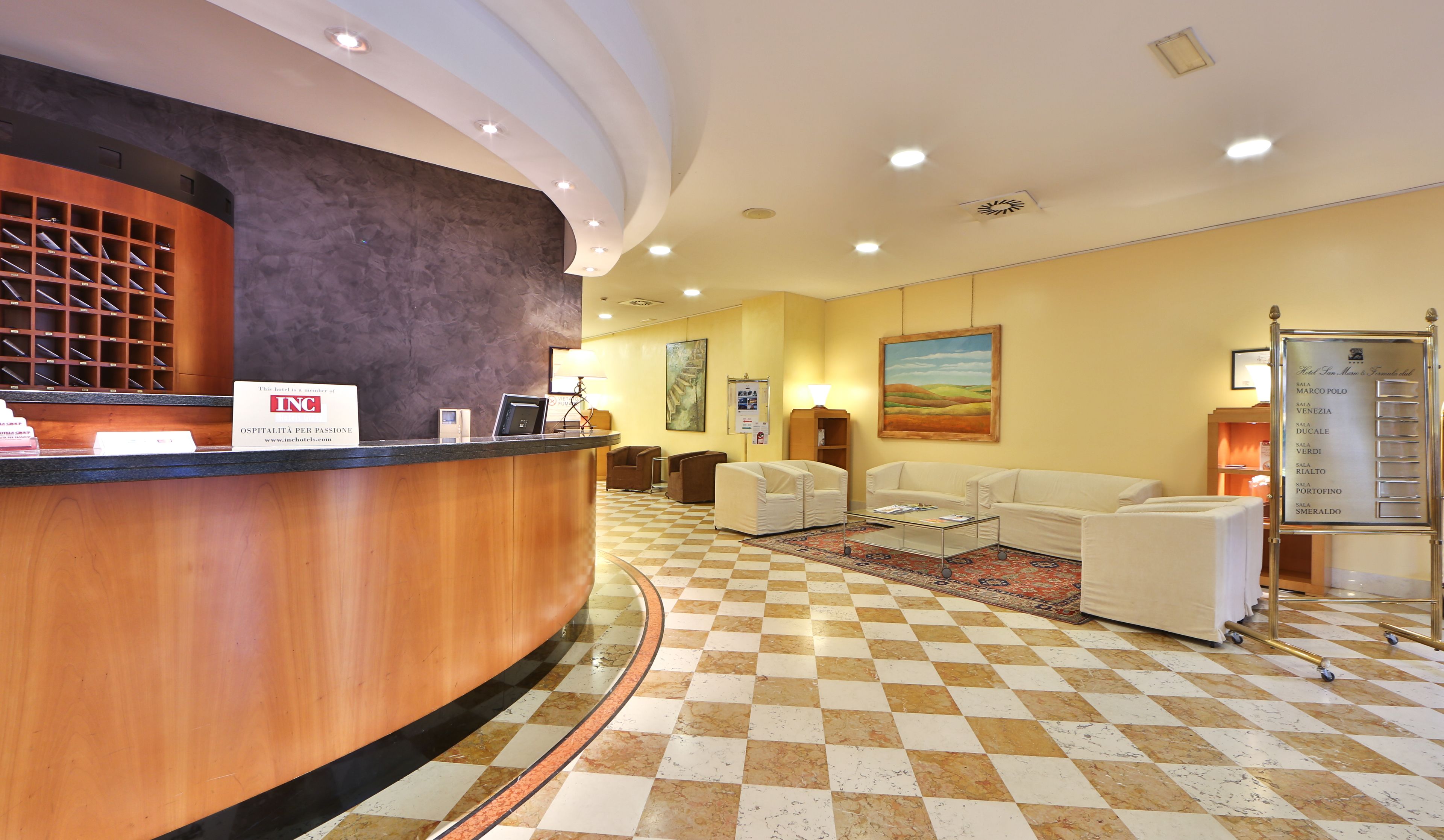 A hotel reception area with a check-in counter, seating area, and a wall-mounted information board listing room types.