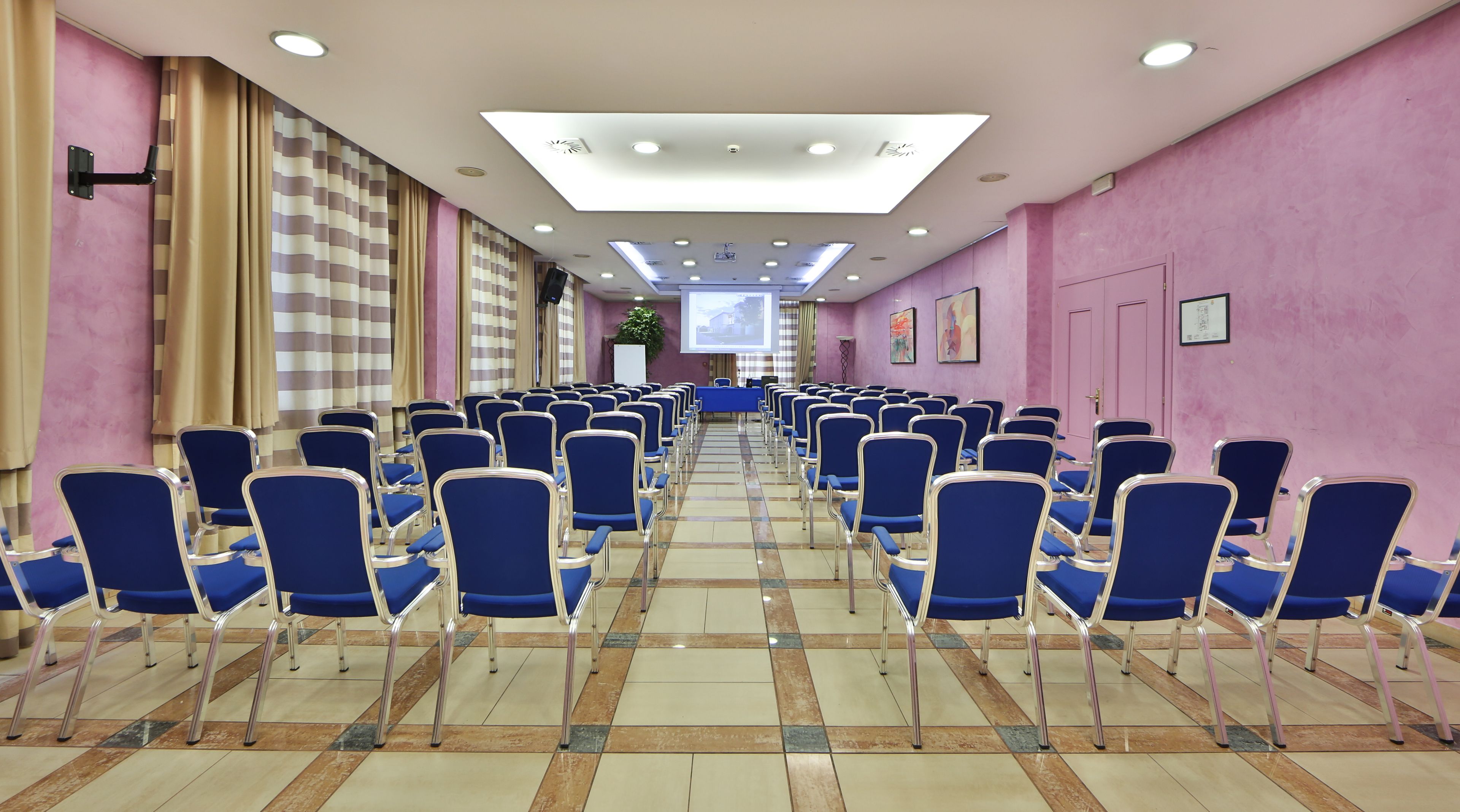 Een vergaderzaal met rijen blauwe stoelen, een projectiescherm en een betegelde vloer.
