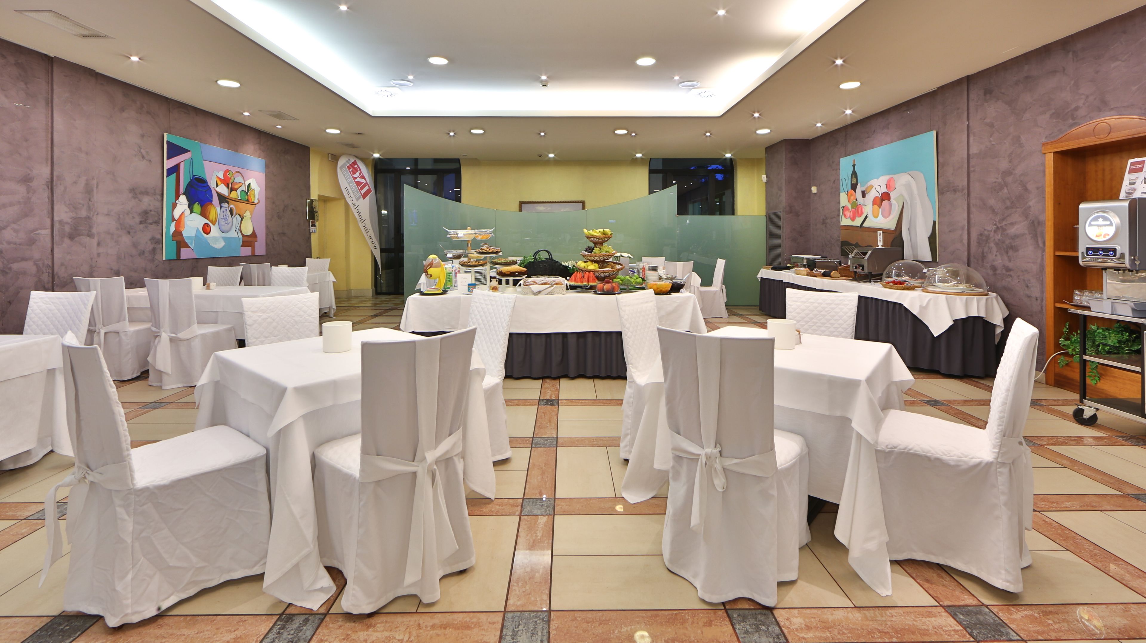 Un salón de banquetes con mesas redondas cubiertas con manteles blancos y sillas, una zona de buffet con comida y bebidas, y obras de arte en las paredes.