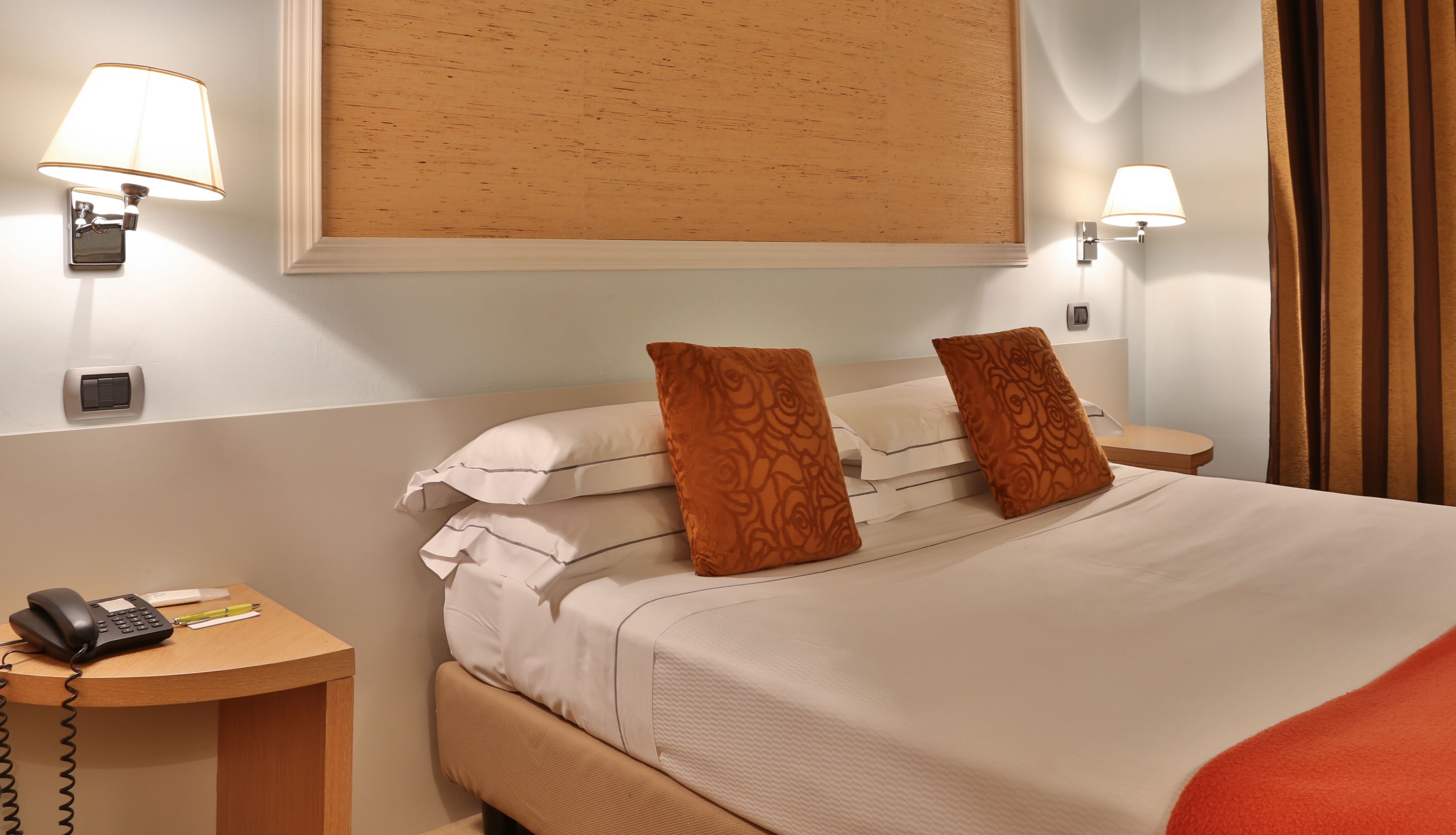 A hotel room with a bed, two pillows, a bedside table with a phone, and two wall-mounted lamps.