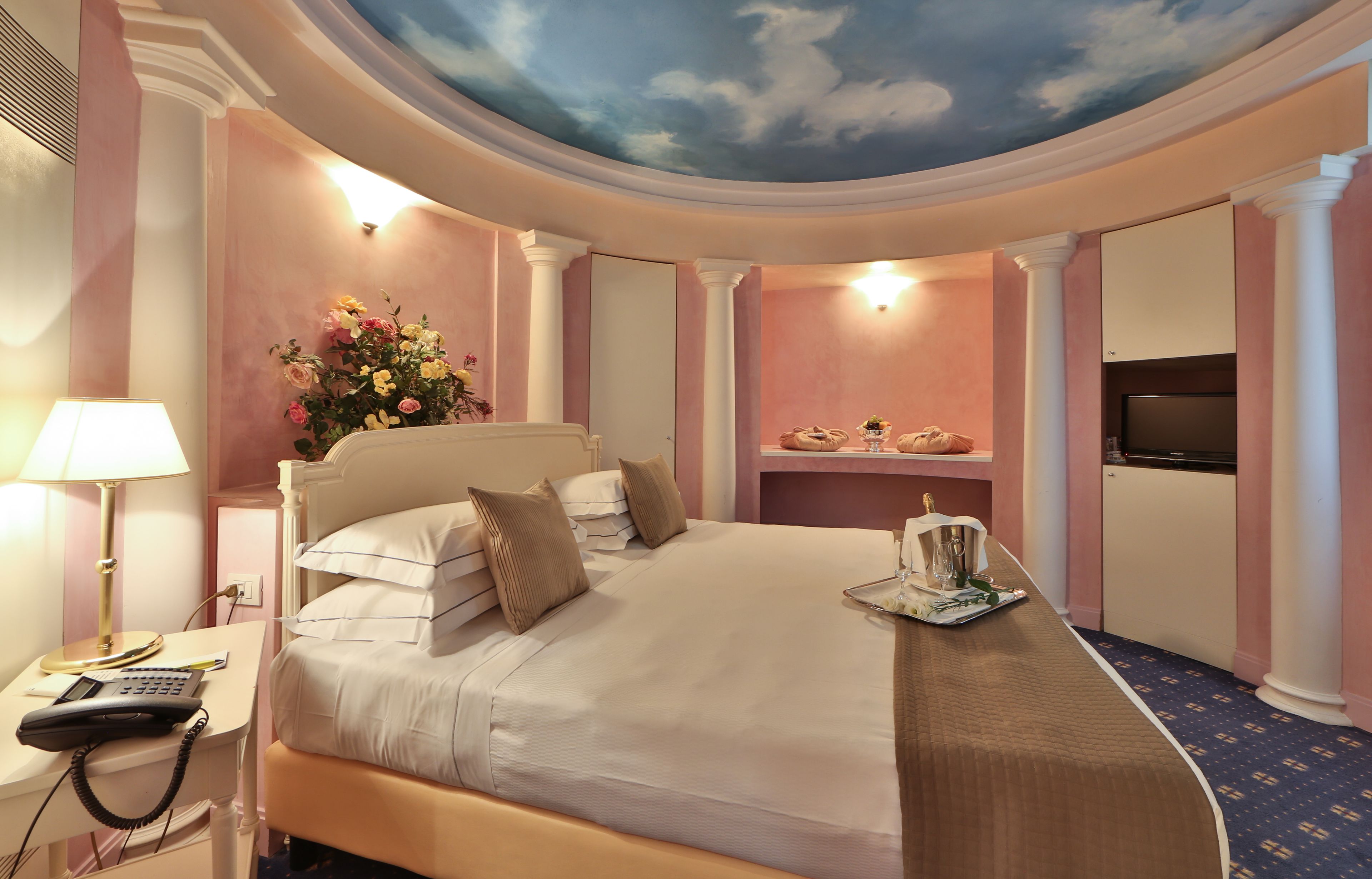 A hotel room with a large bed, a desk with a lamp, a telephone, a television, and a decorative ceiling mural.