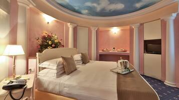 Habitación de hotel con una cama grande, un escritorio con lámpara, teléfono, televisión y un mural decorativo en el techo.