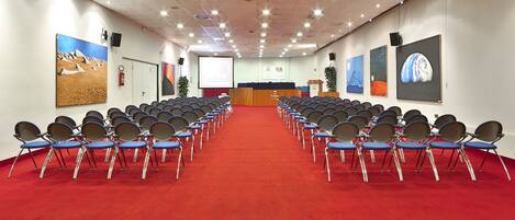 Un salón de conferencias con filas de sillas, un atril y una pantalla al frente.