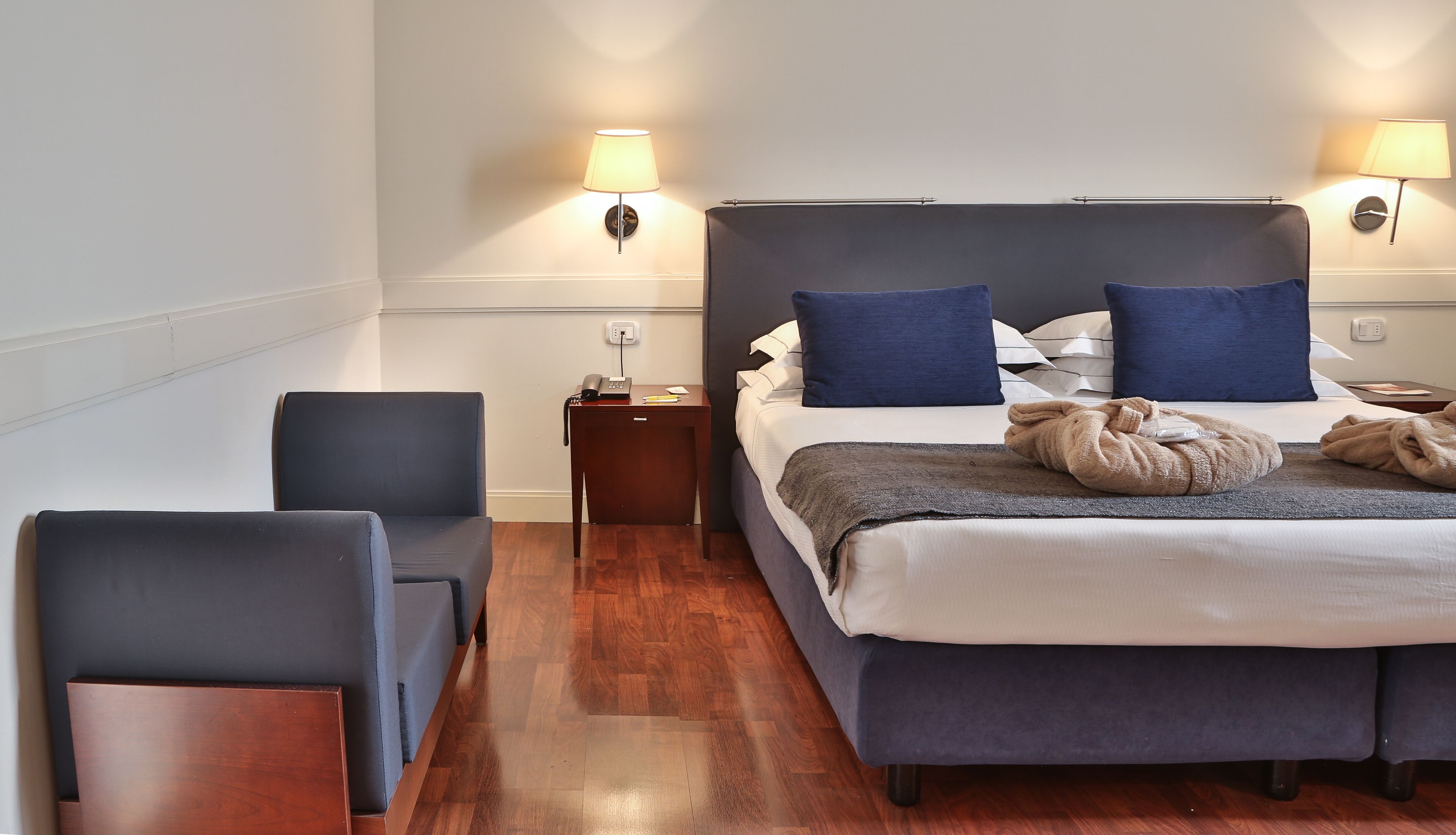 Une chambre d’hôtel avec un lit, deux lampes de chevet, une table de chevet avec un téléphone, une chaise et un plancher en bois.