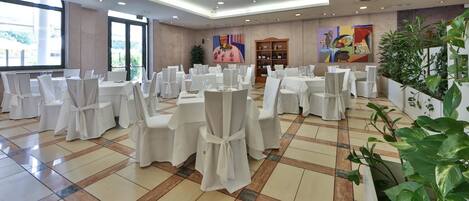 A banquet hall with round tables covered in white tablecloths and chairs, a wine rack, and large windows with a view of greenery.