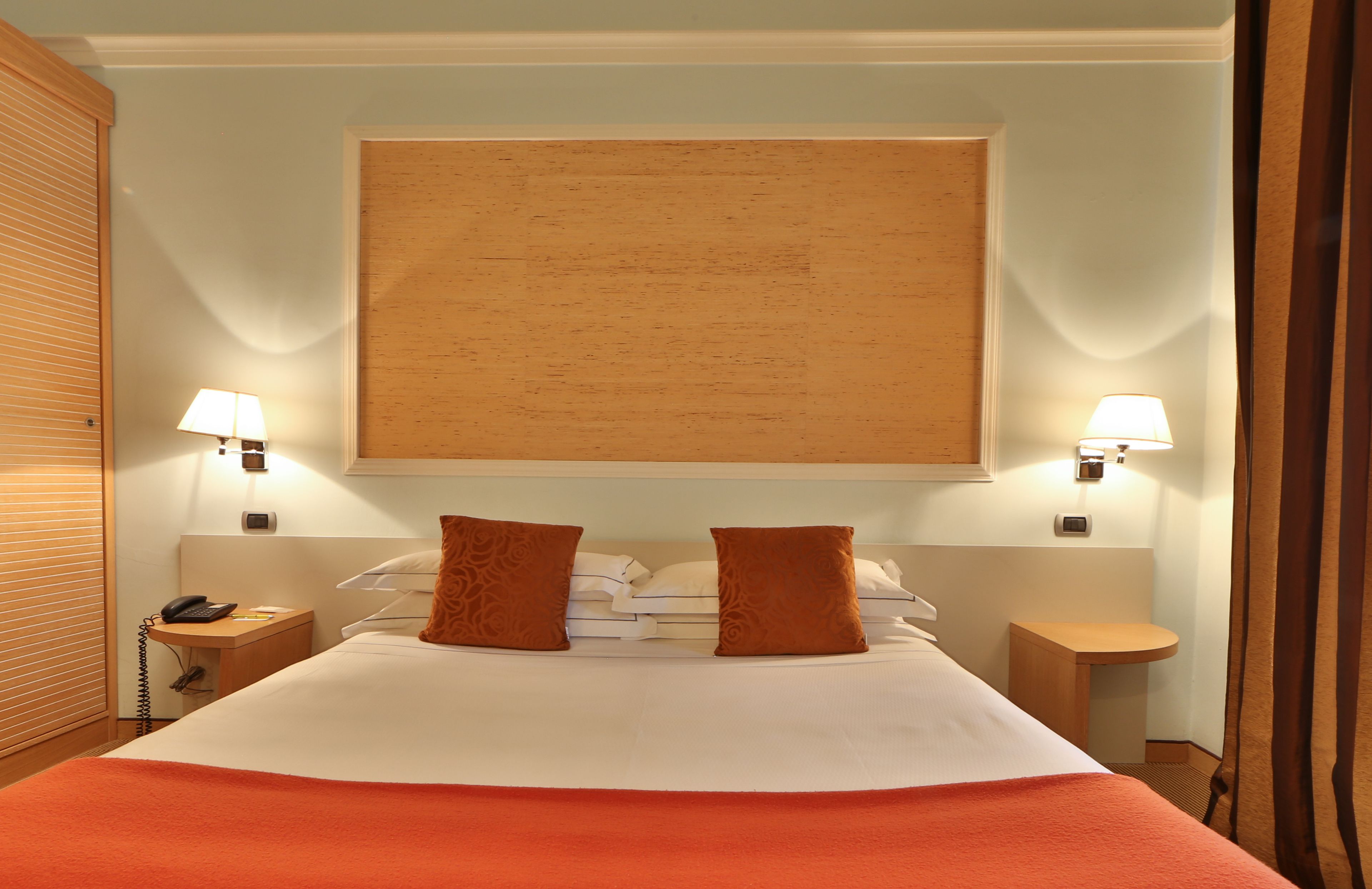 A hotel room with a large bed, two bedside lamps, a nightstand with a phone, and a wooden headboard.