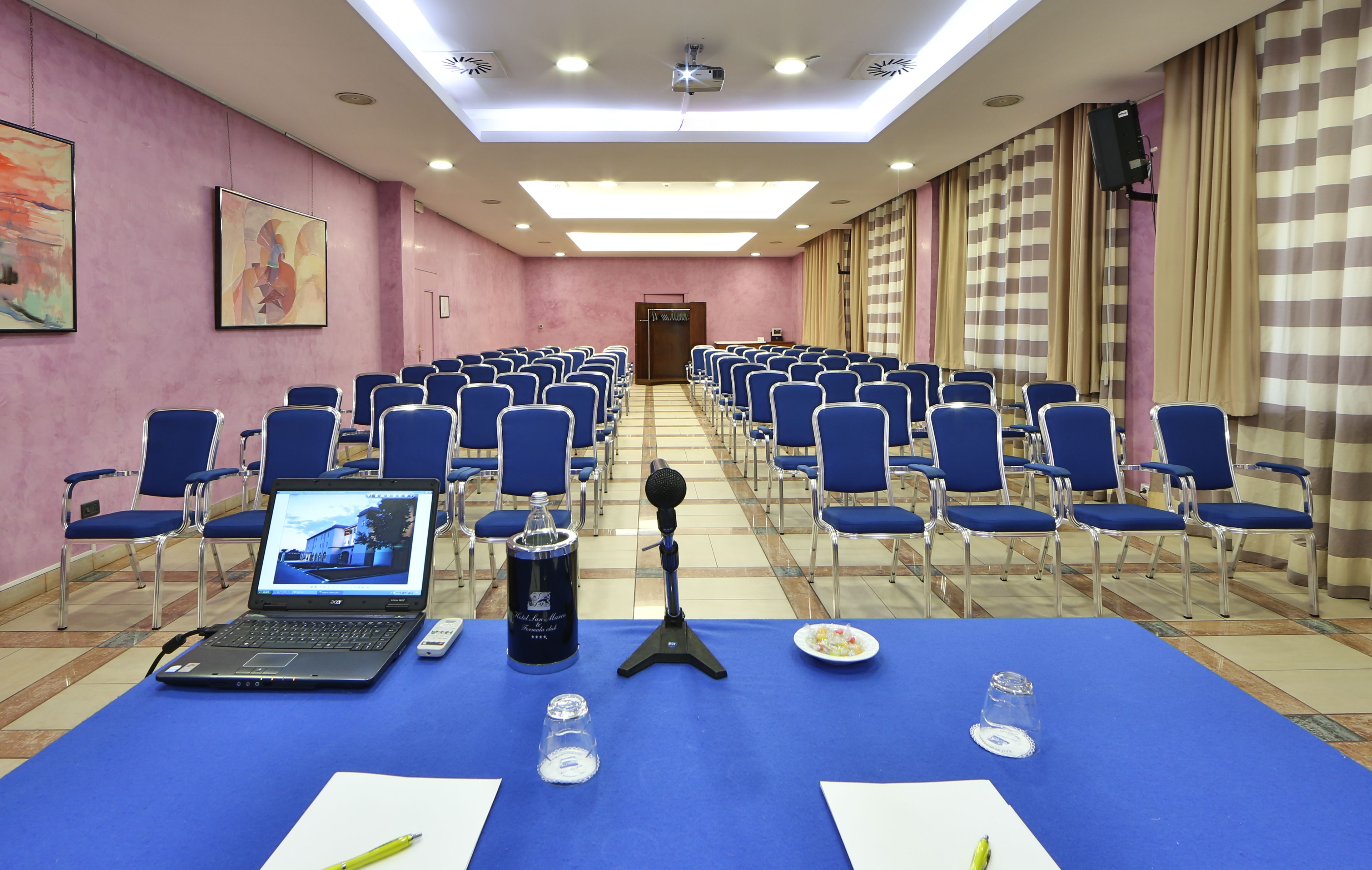 Una sala conferenze con file di sedie blu, un portatile su un tavolo, un microfono e un proiettore a soffitto.