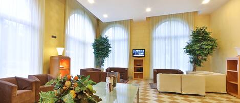 A hotel lobby with a checkered floor, two large potted plants, a flat-screen TV mounted on the wall, and multiple seating areas including sofas and armchairs.