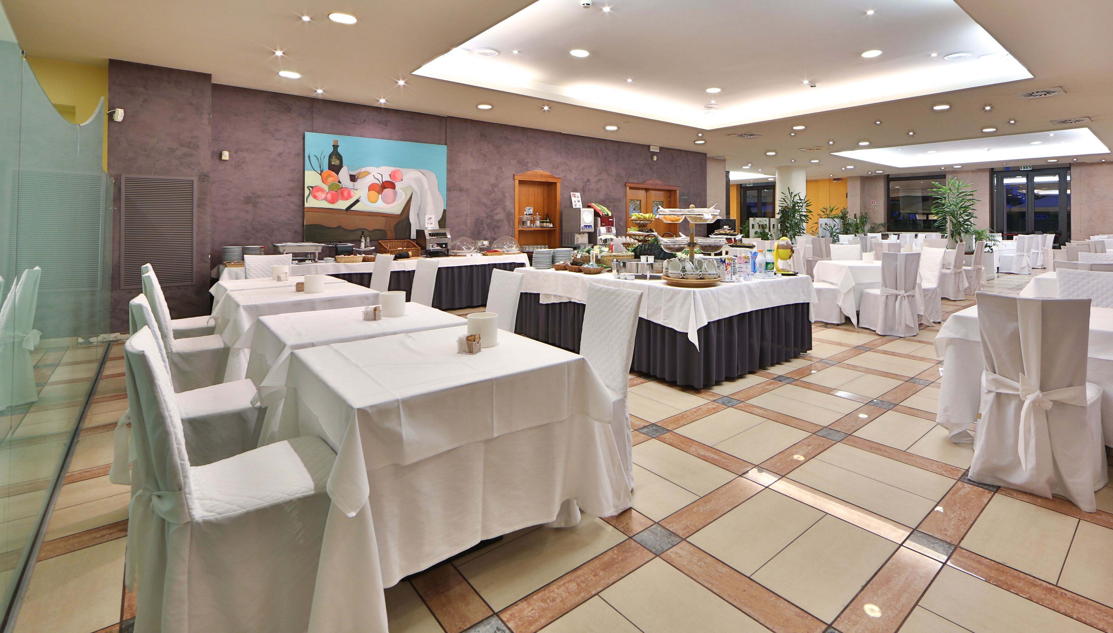 A banquet hall with multiple tables set for dining, a large painting on the wall, and a ceiling with recessed lighting.