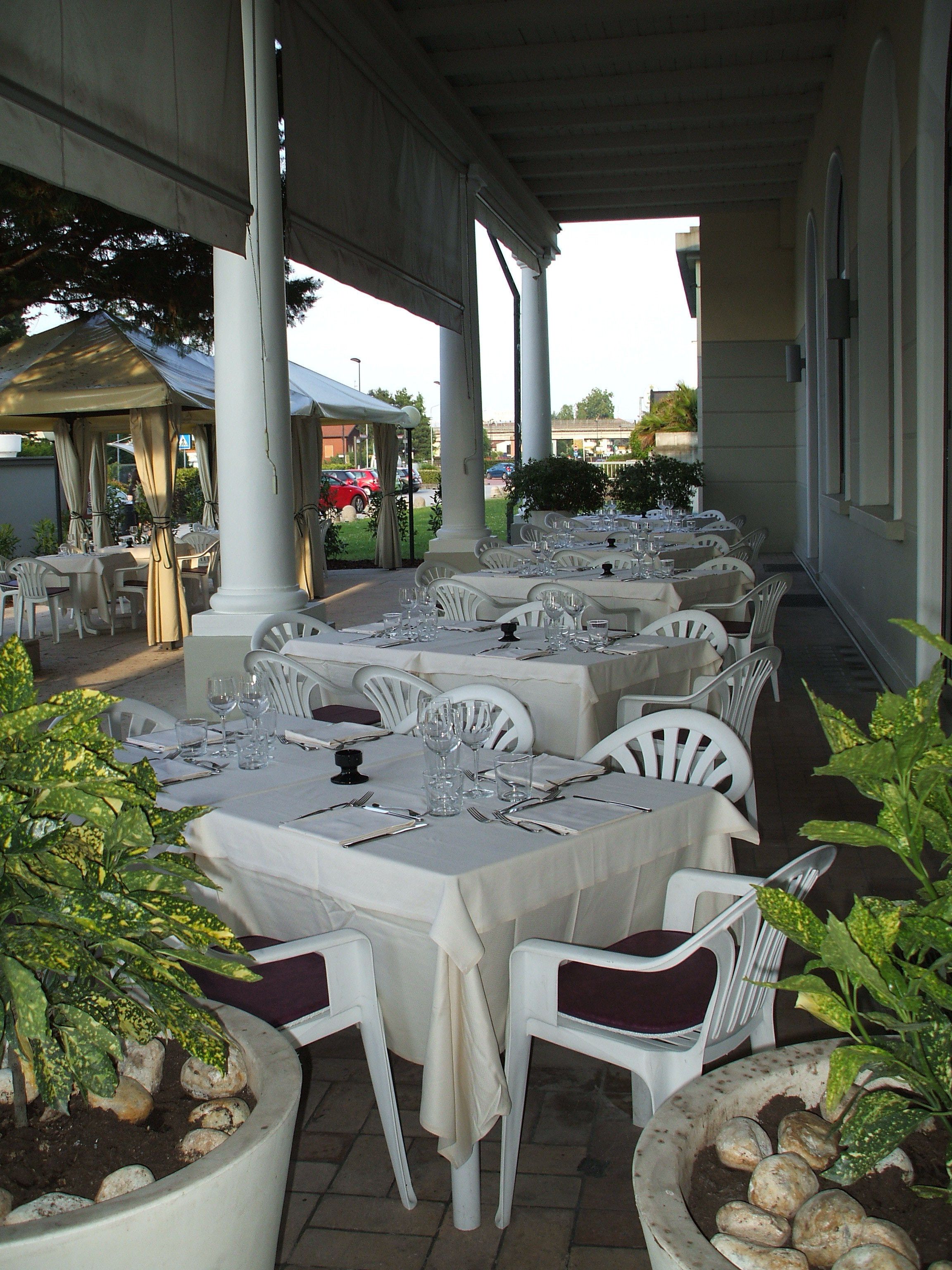 A covered outdoor dining area with tables set for a meal, surrounded by white chairs and potted plants.