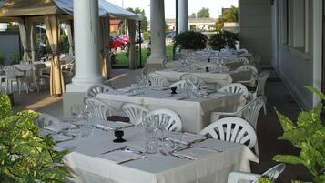 A covered outdoor dining area with tables set for a meal, surrounded by white chairs and potted plants.